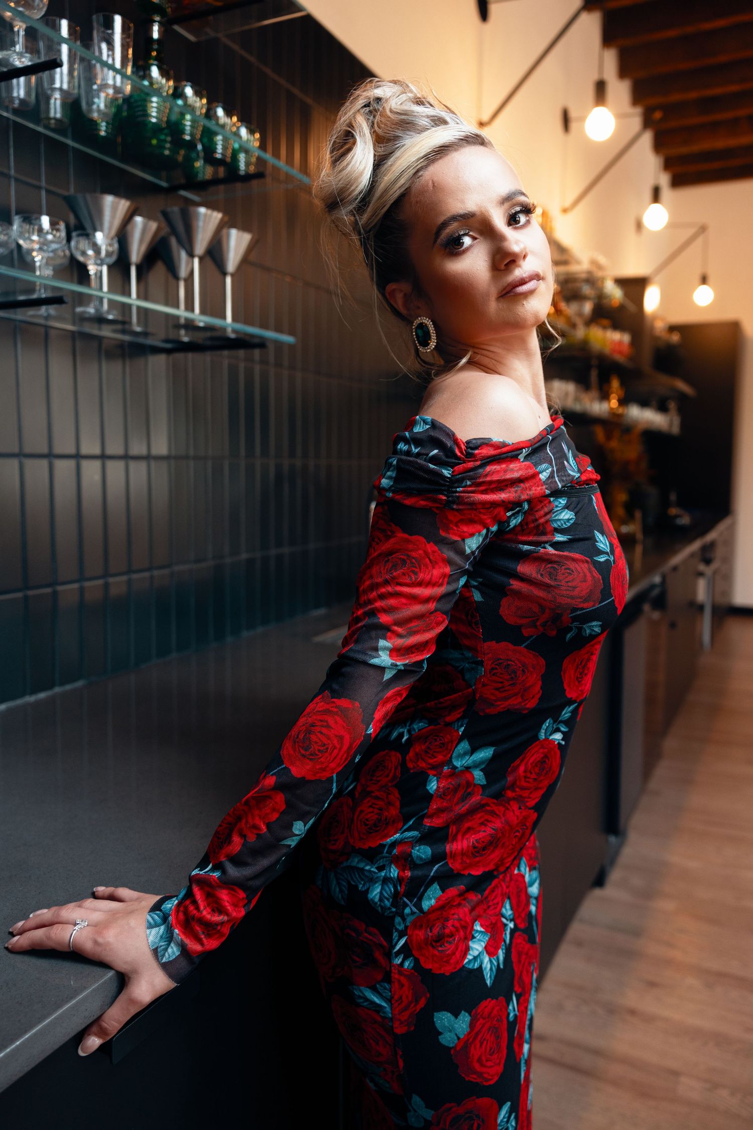 Woman in floral red rose dress posing elegantly at a dark bar counter.