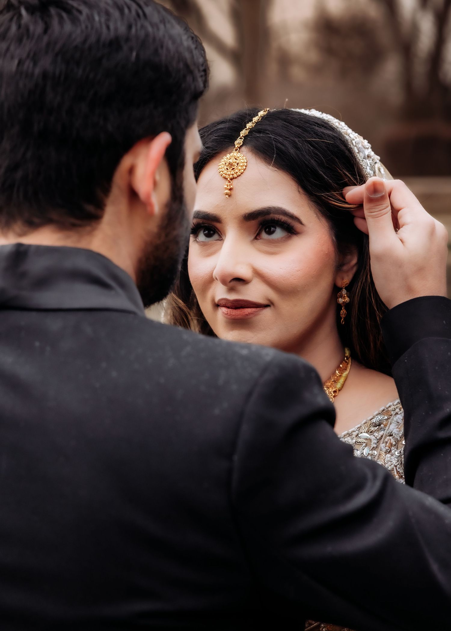 Groom adjusts bride's maang tikka jewelry as she gazes up adoringly wearing traditional South Asian bridal look.