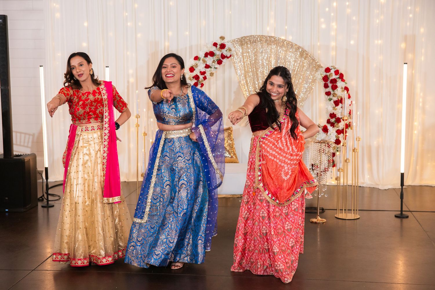 Three women perform a vibrant Bollywood dance in colorful lehengas at an Indian wedding reception.