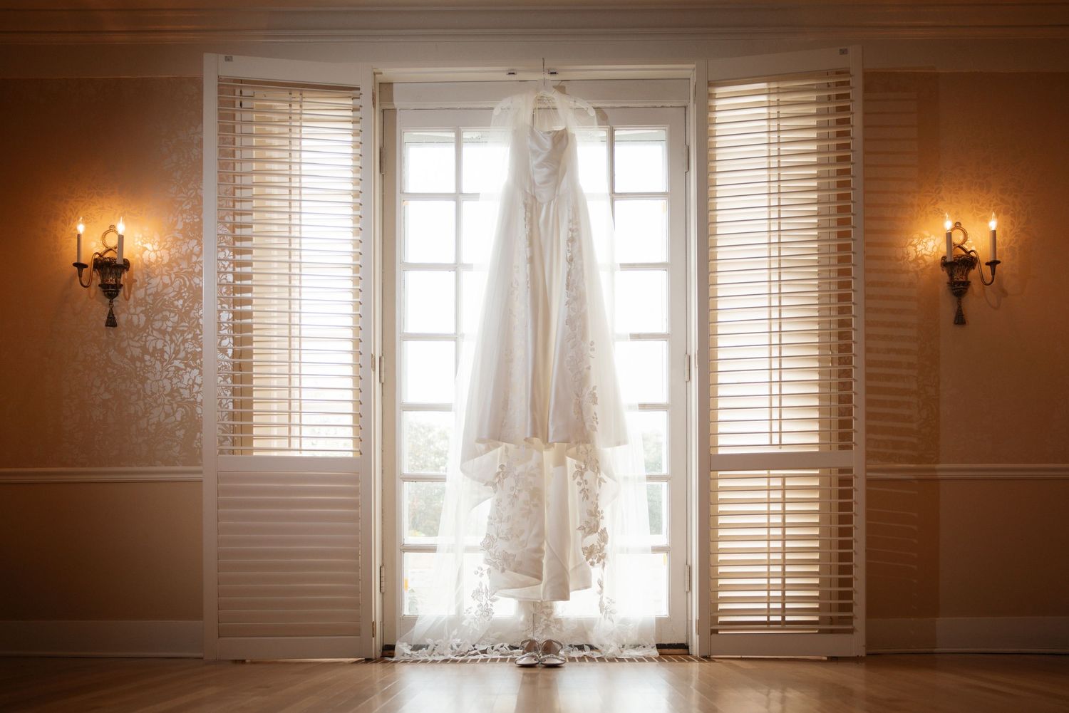 Elegant lace wedding dress hanging between French doors with shutters and candlelit wall sconces.
