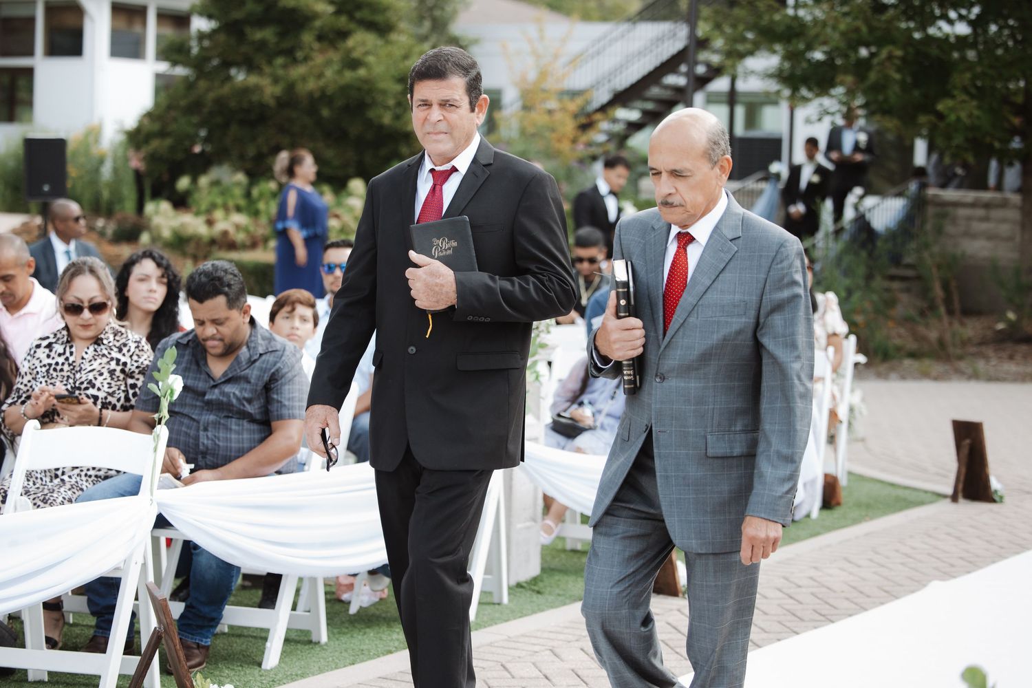 Two men in suits walk down wedding aisle as guests watch the outdoor ceremony.