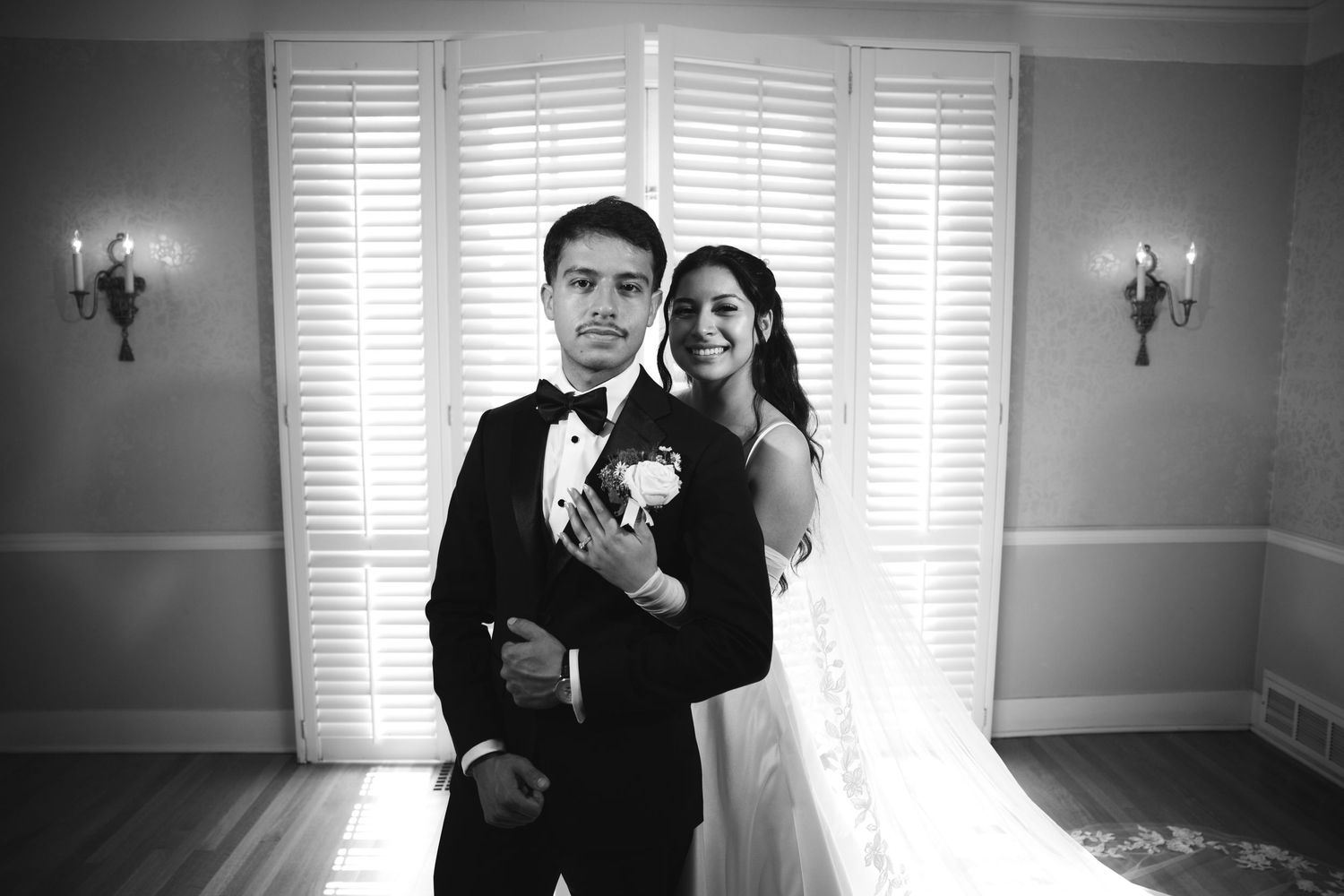 Black and white portrait of bride and groom embracing tenderly in front of shuttered white doors.