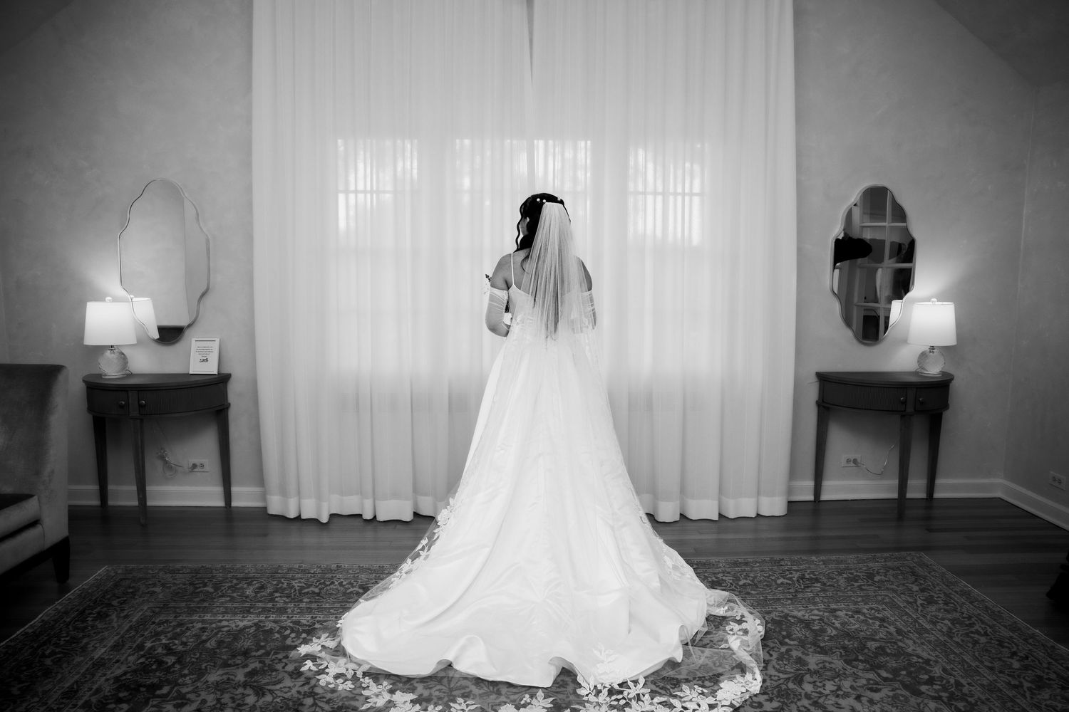 Elegant black and white portrait of bride in ball gown with cathedral veil standing before draped curtains.