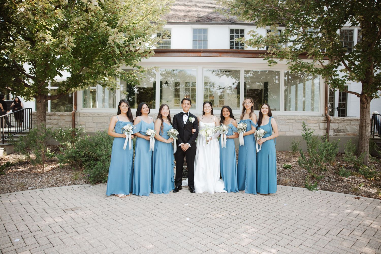 Wedding party portrait with bride, groom, and bridesmaids in blue dresses before a white building.