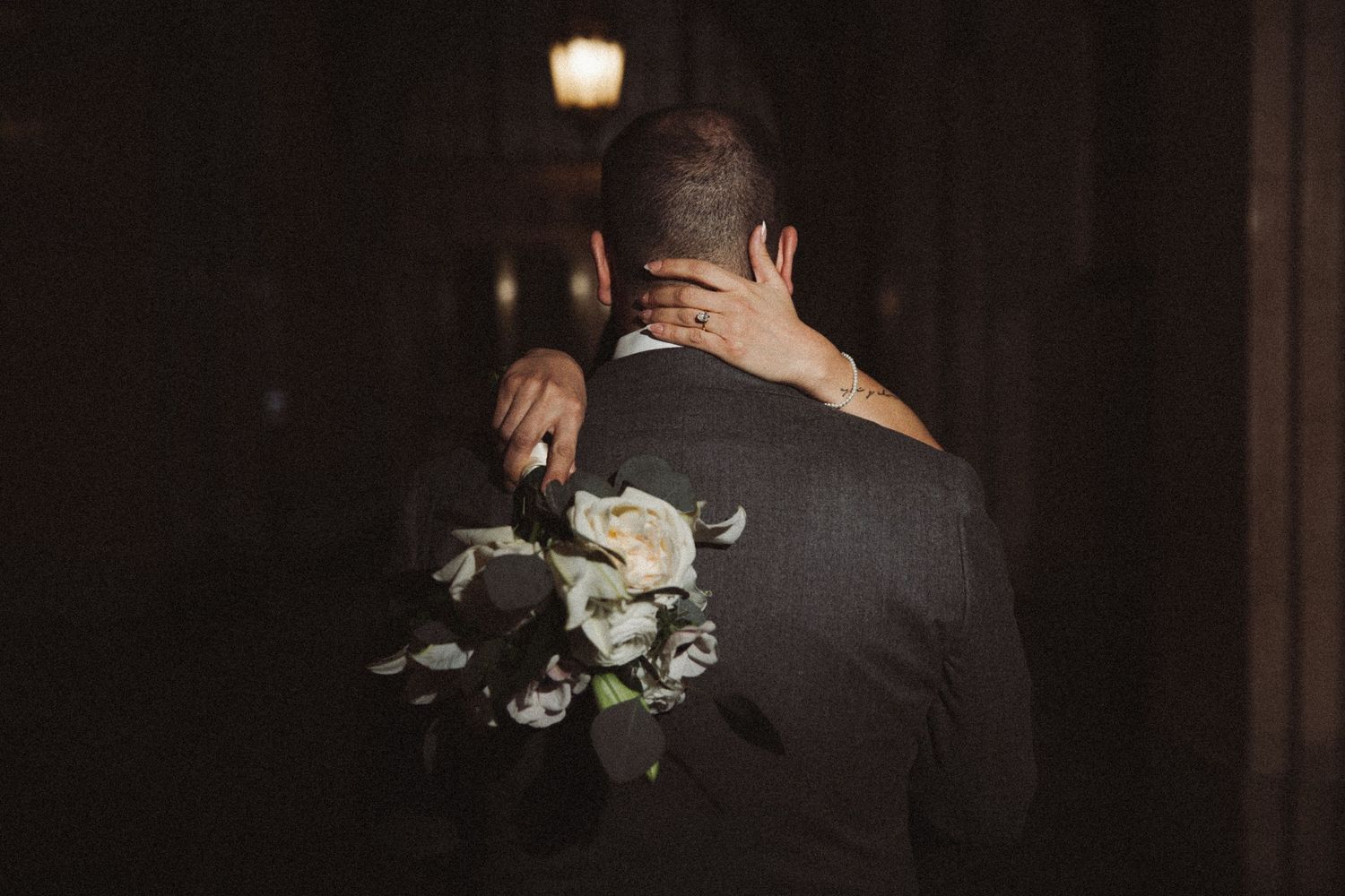 Groom holding white rose bouquet with bride embracing him from behind in dim light.