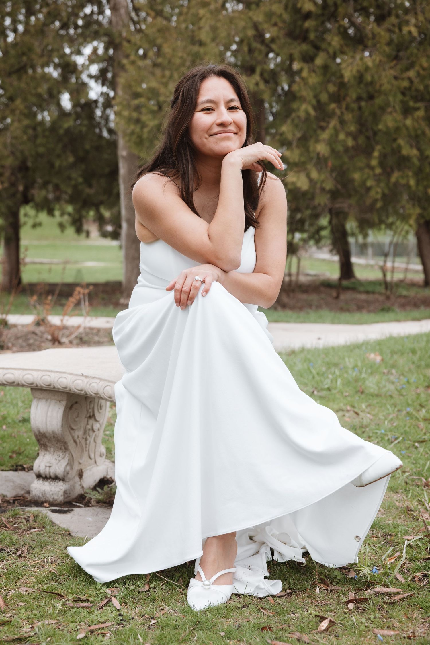 Young woman in white dress sitting on stone bench in park, smiling gracefully with hand on chin.
