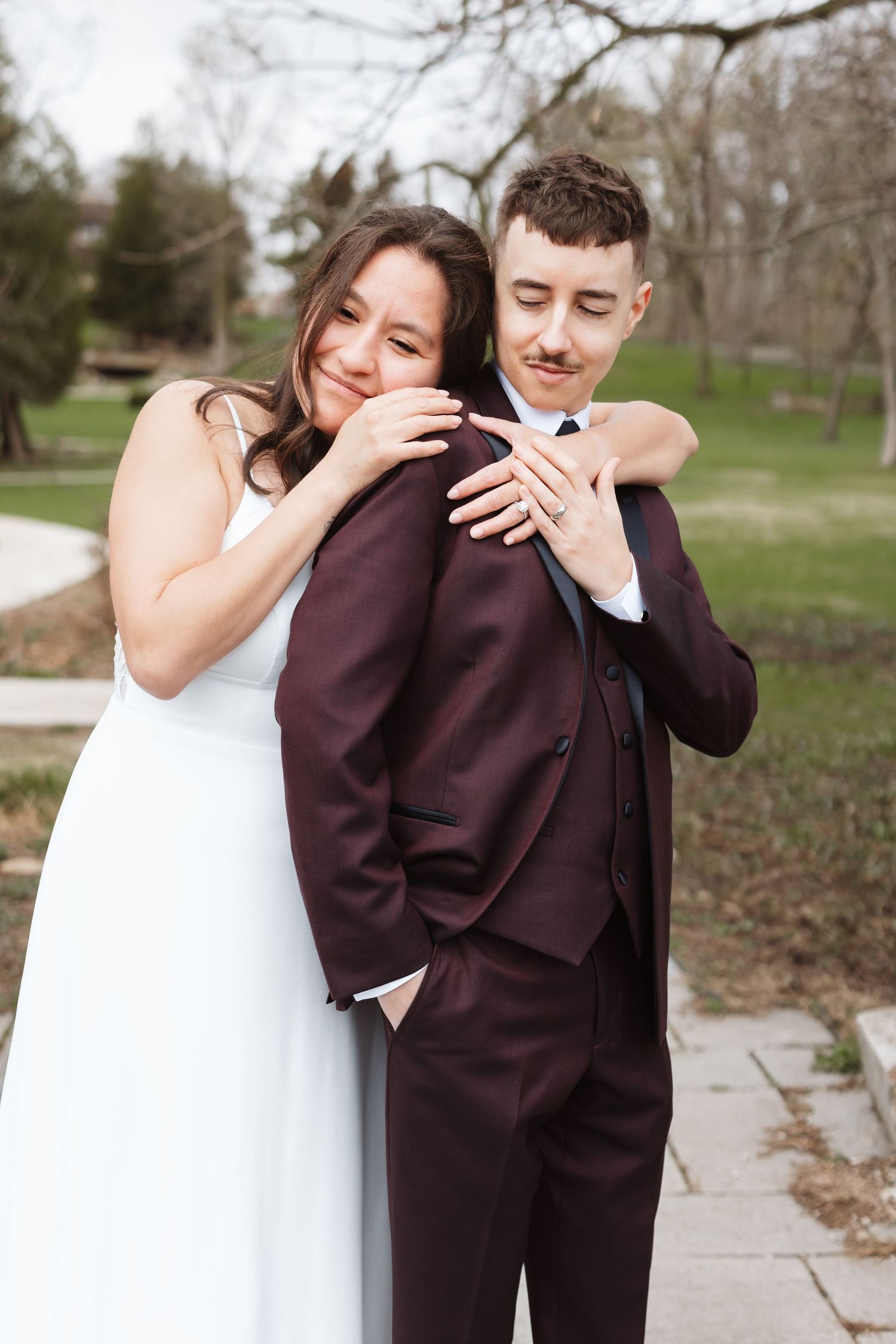 Bride in white dress embraces groom in burgundy suit outdoors, both smiling in a romantic wedding portrait.