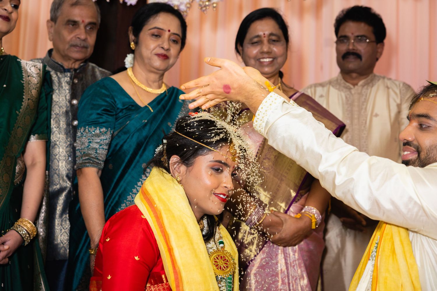 Indian wedding ritual with groom pouring yellow rice on brides head surrounded by smiling family members.