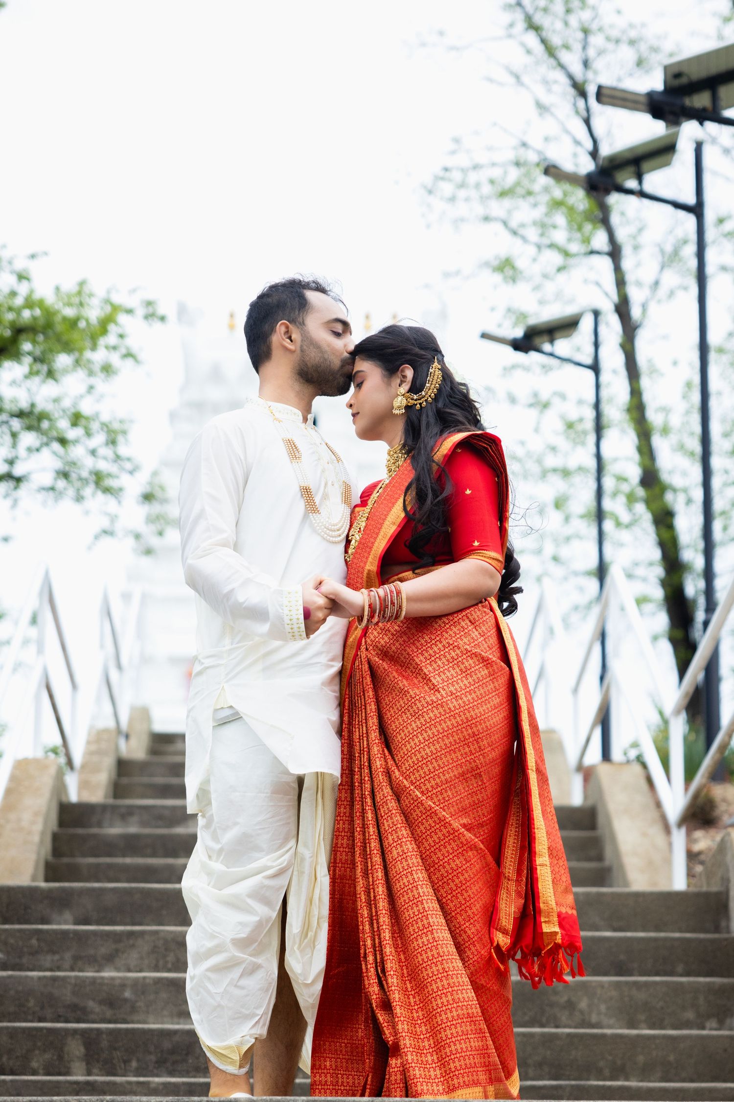 Indian bride in red saree and groom in white kurta share a romantic kiss on outdoor staircase.
