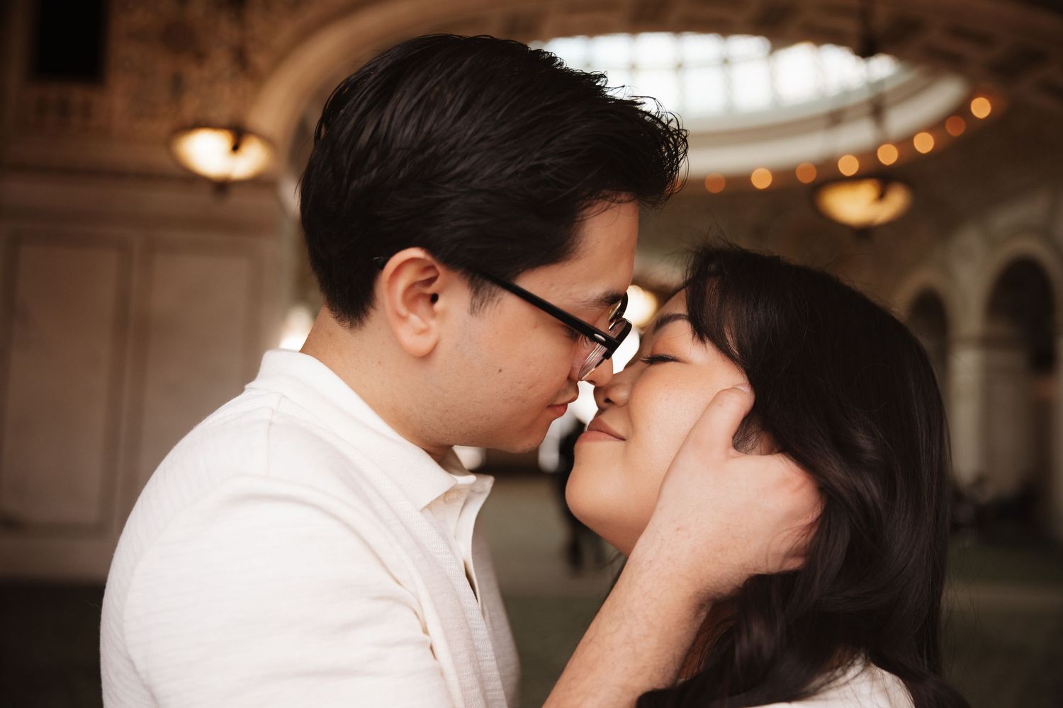 Romantic couple sharing a gentle kiss in elegant domed interior with warm glowing chandelier lights above.