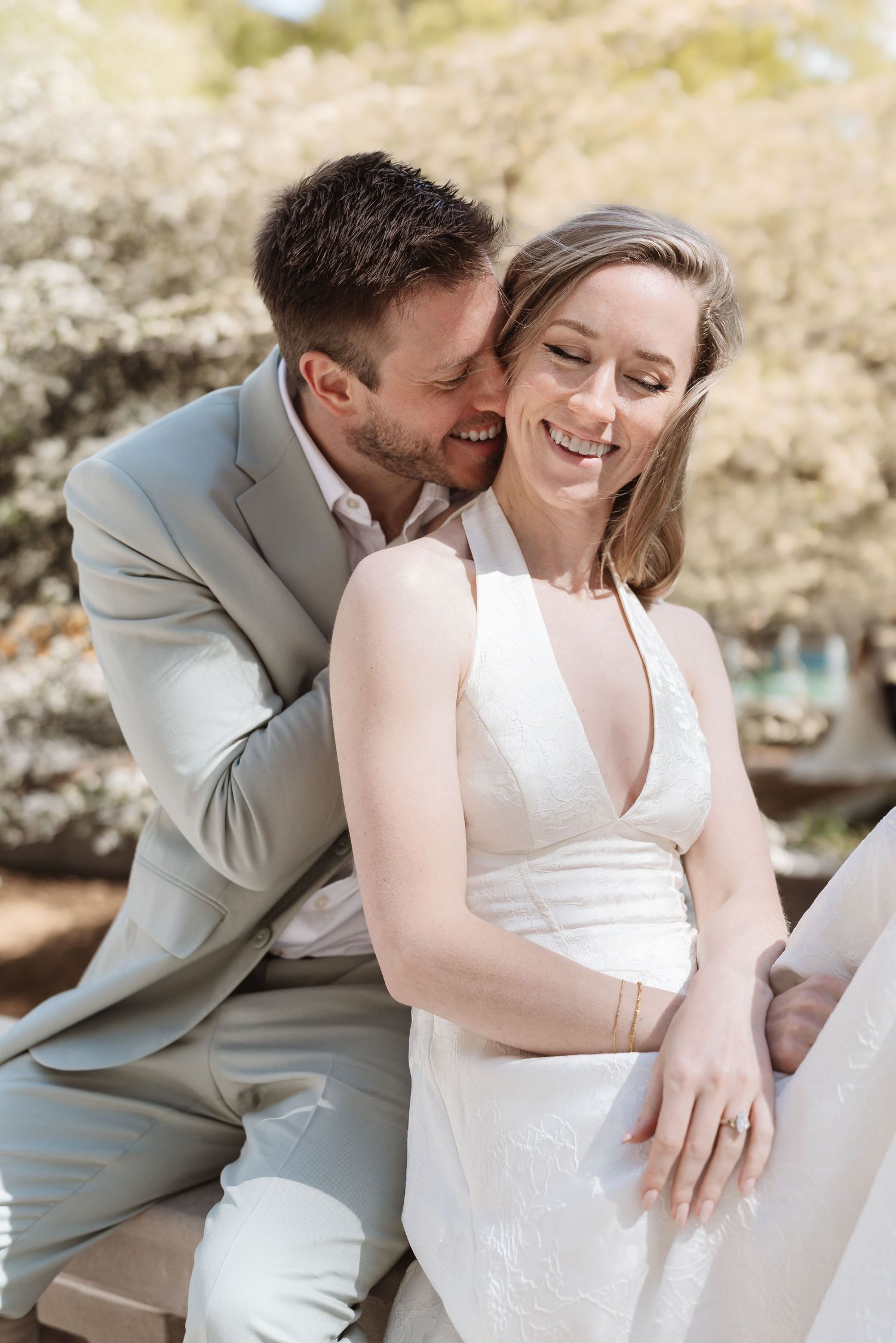 Laughing bride in white dress and groom in gray suit share a joyful moment on an outdoor bench.