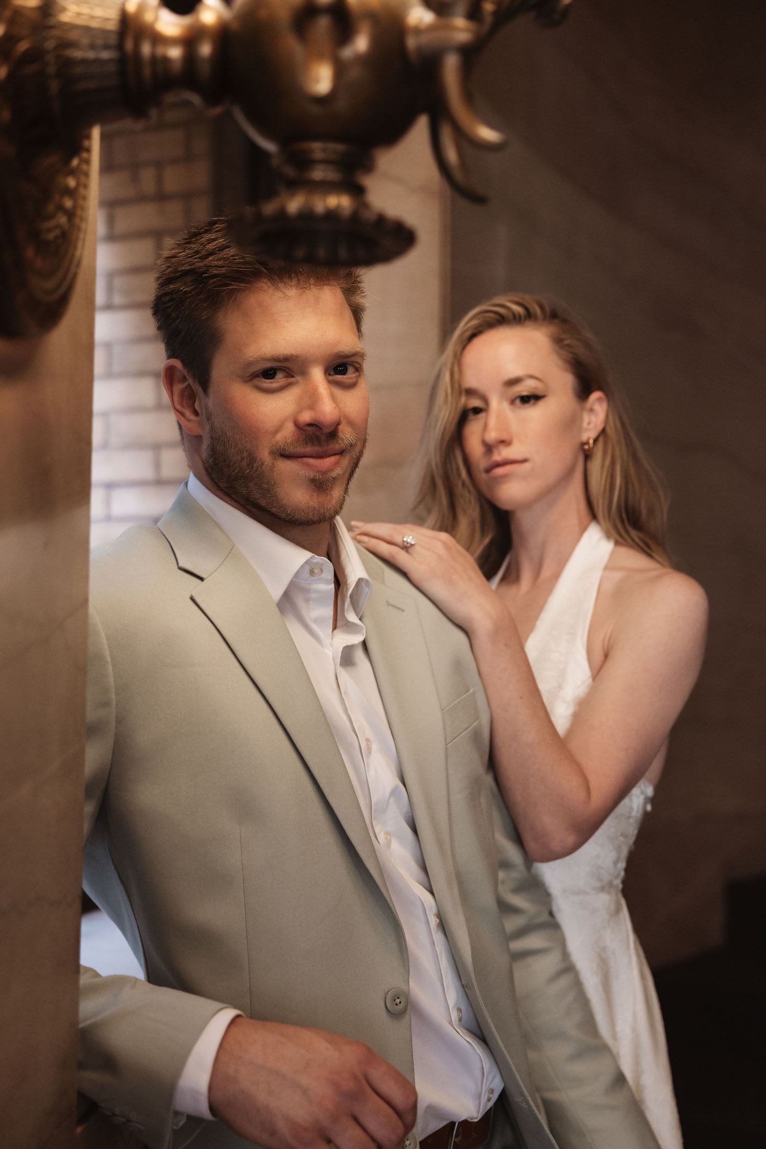 Groom in light suit smiles at camera while bride in white dress stands behind him touching his shoulder.