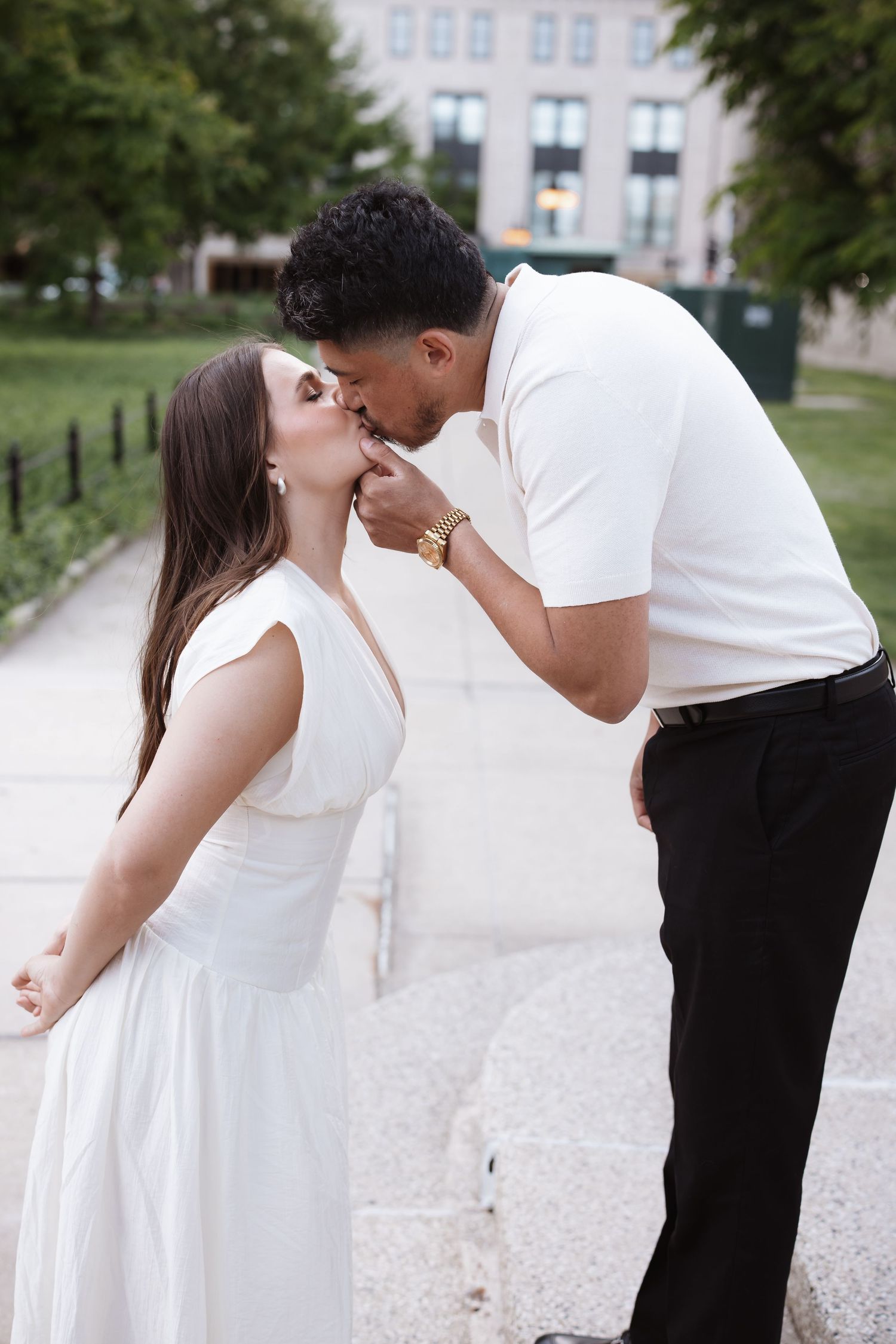 Couple shares a tender kiss outdoors, man in white polo and gold watch, woman in elegant white dress.