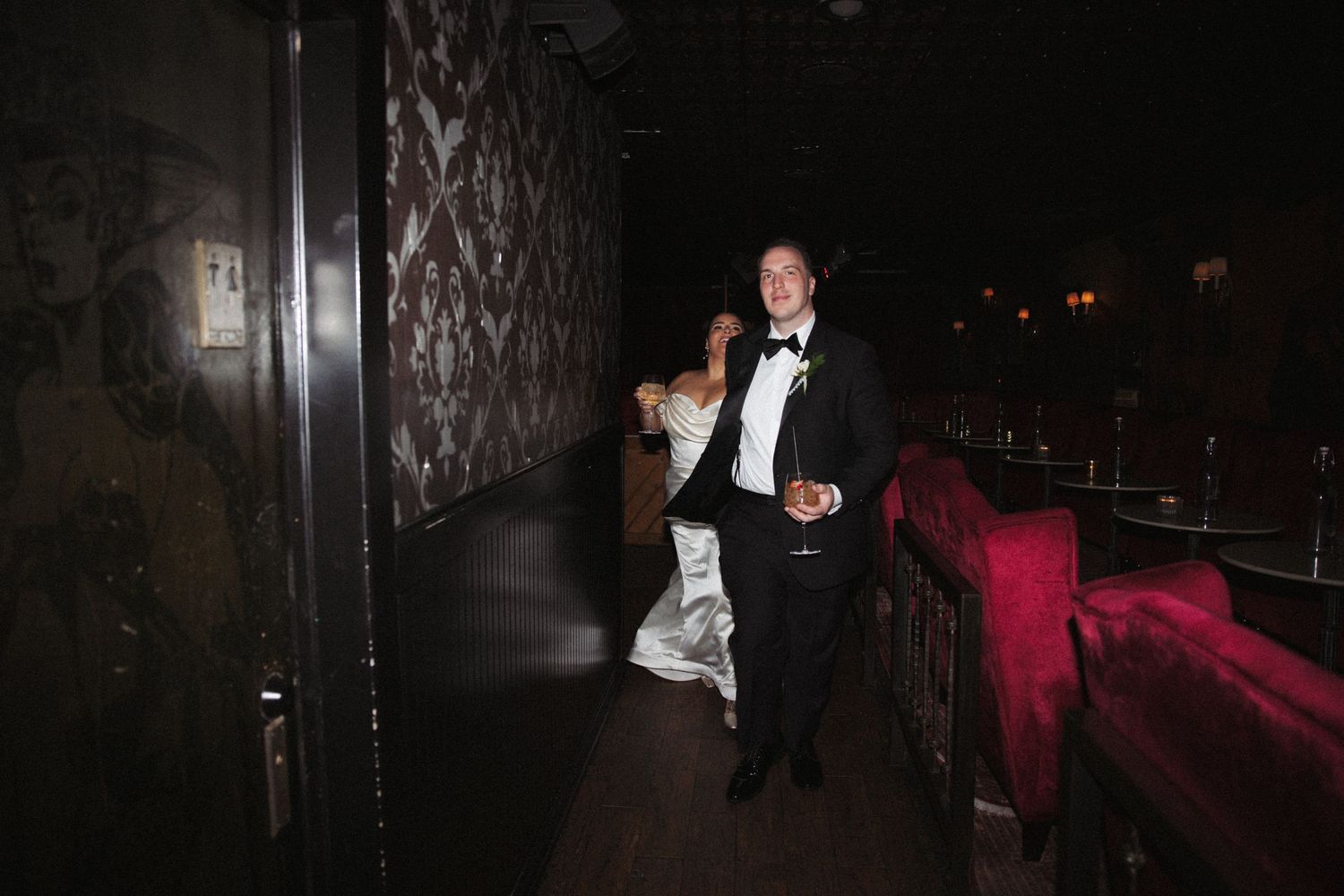 Bride in mermaid gown and groom in tuxedo walking through dramatic dark corridor at wedding reception venue.