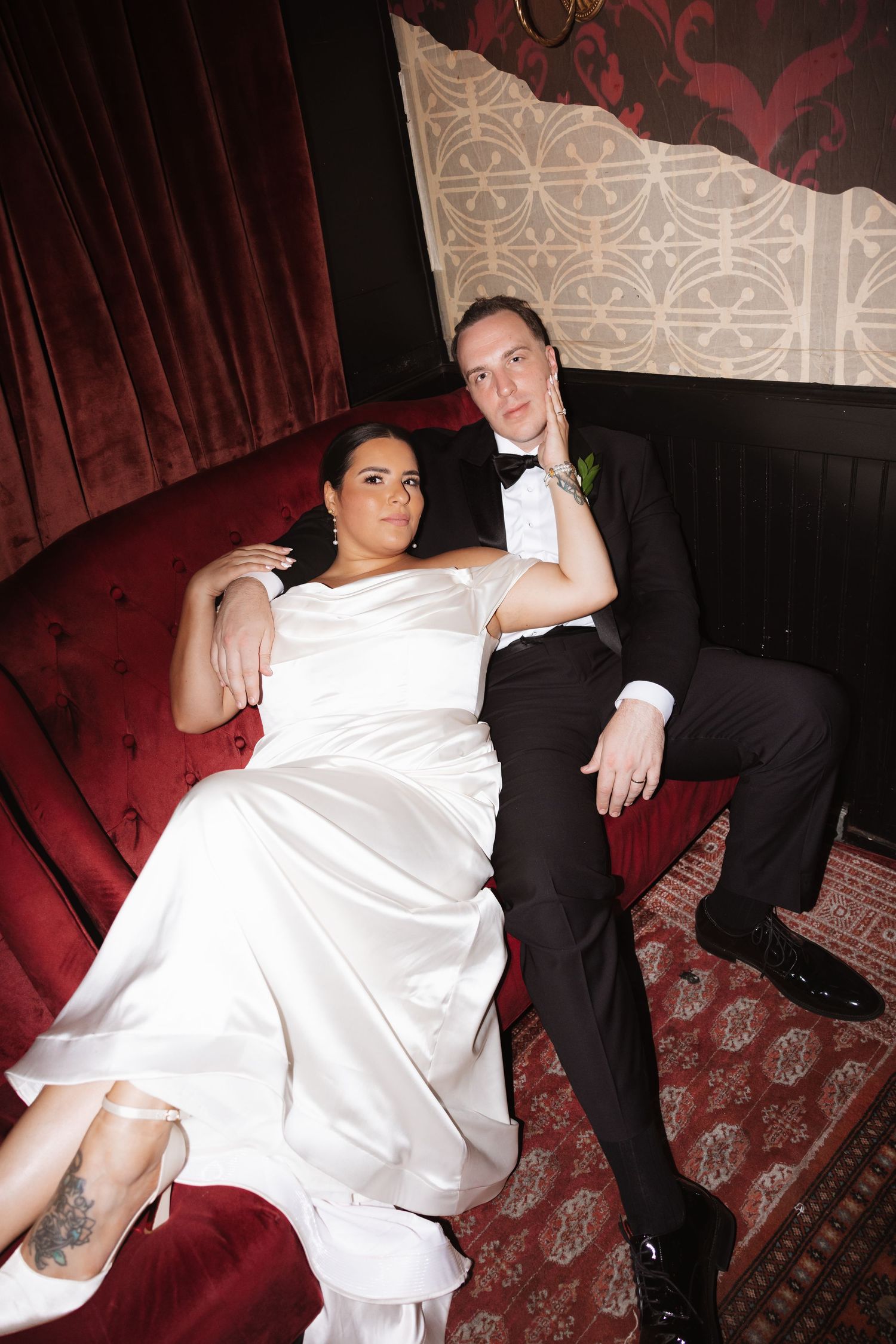 Bride and groom relaxing on a red velvet sofa, wearing wedding attire, in an elegant dark reception venue.