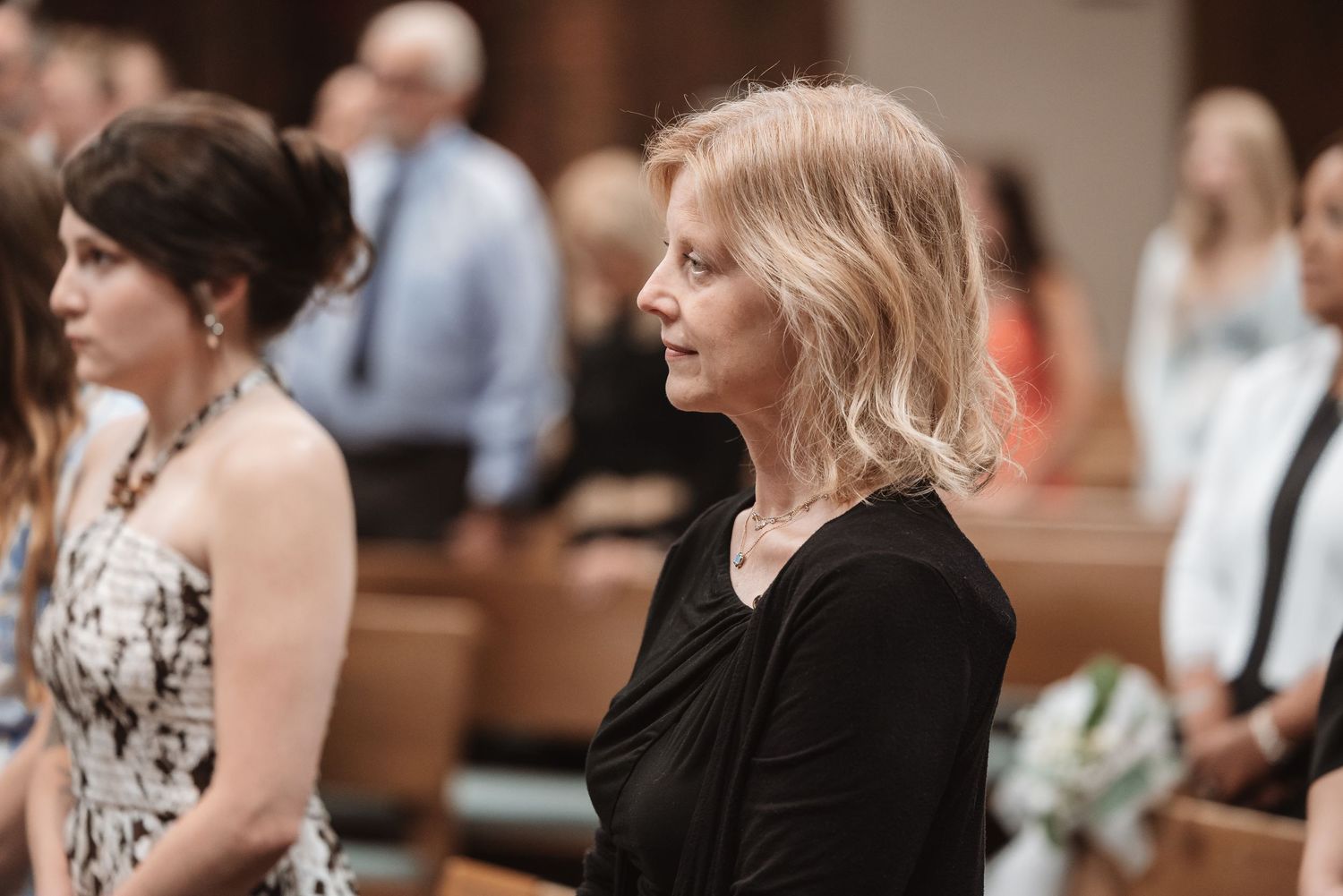Guests standing in church pews during a wedding ceremony, woman in black dress in foreground.