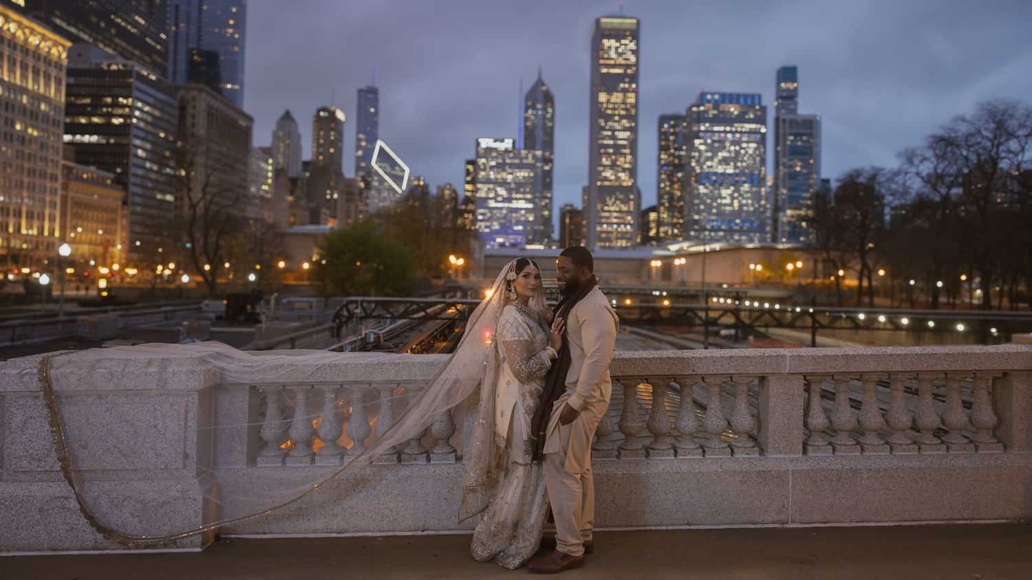 Interracial wedding couple pose on bridge with stunning Chicago skyline illuminated at twilight.