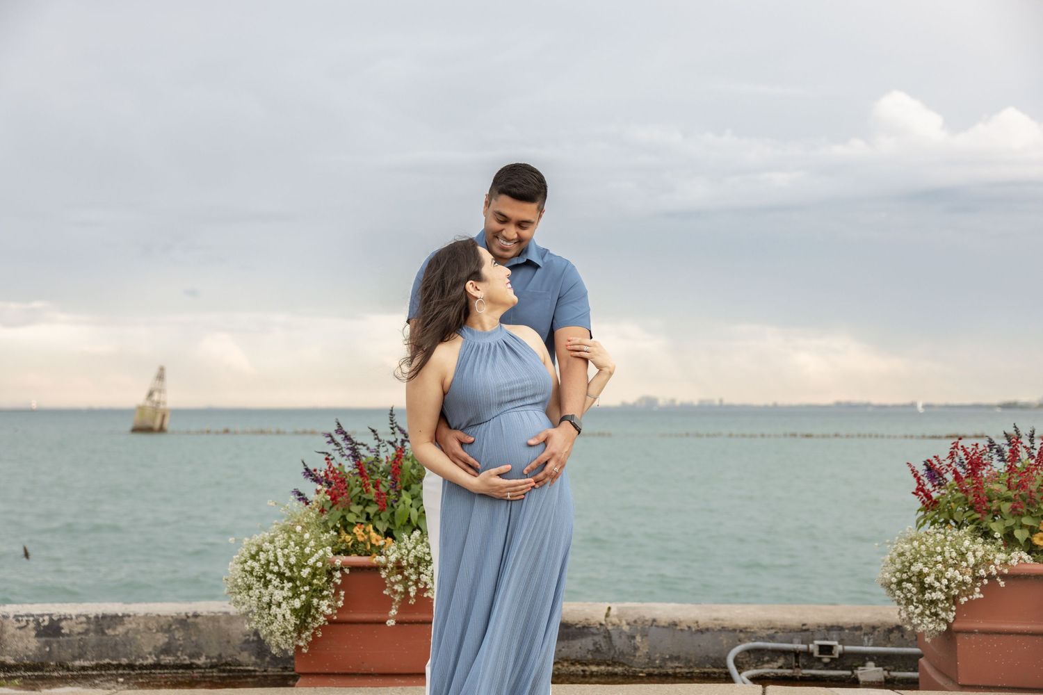 Pregnant couple embracing by the waterfront, woman in blue maxi dress, man in blue shirt, flowers and sailboat in background.