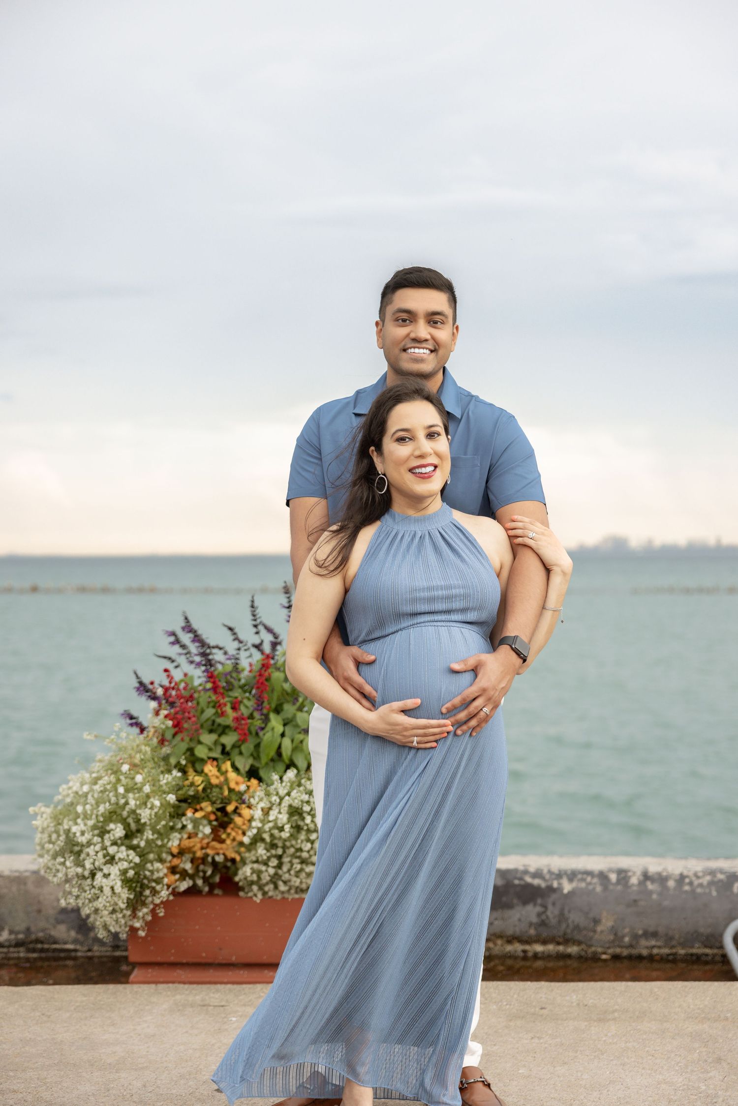 Smiling maternity couple by the lakefront, man in blue polo embracing woman in flowing blue halter dress, flowers nearby.