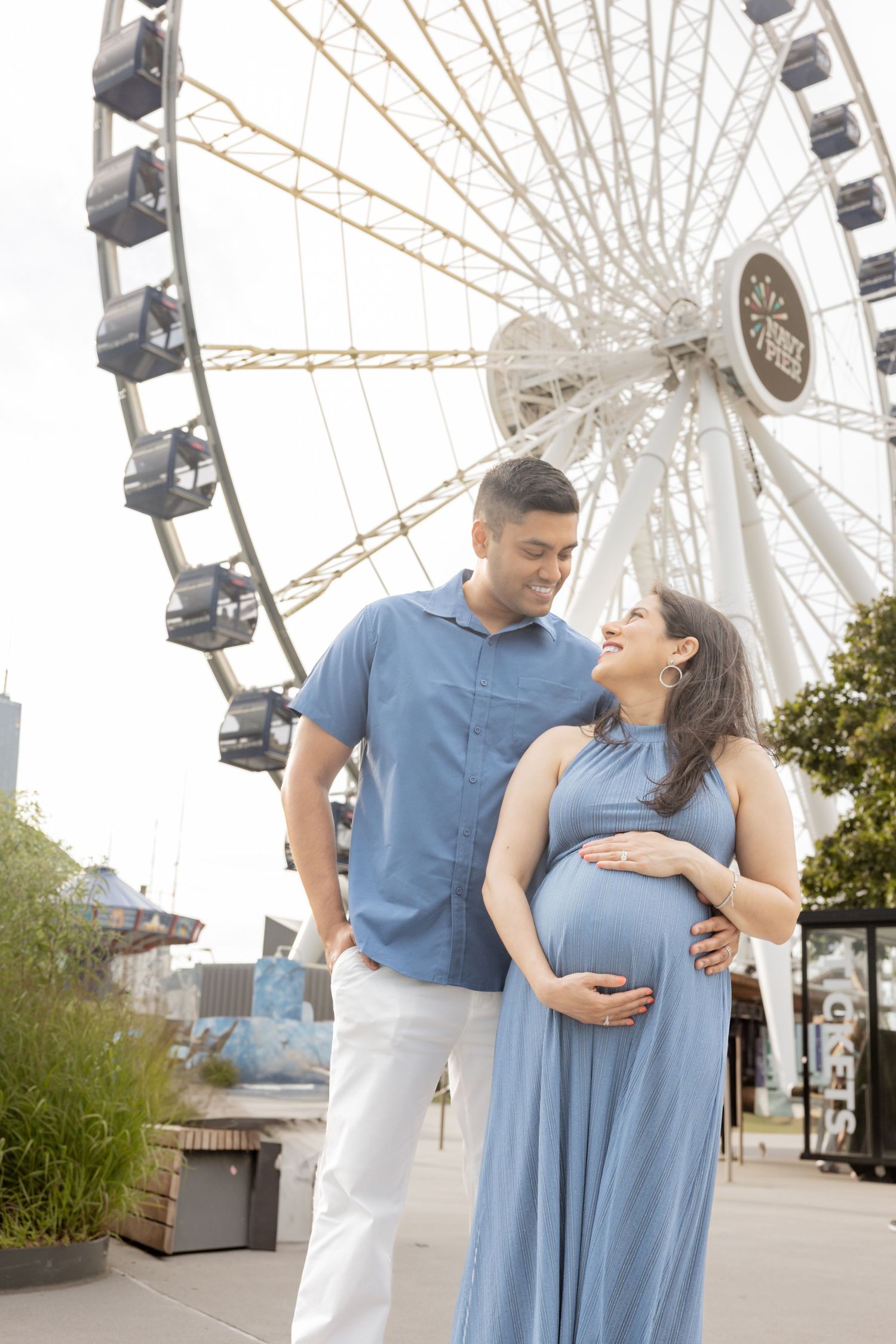 Expecting couple smiling at each other in front of a Ferris wheel, woman in blue dress holding bump.