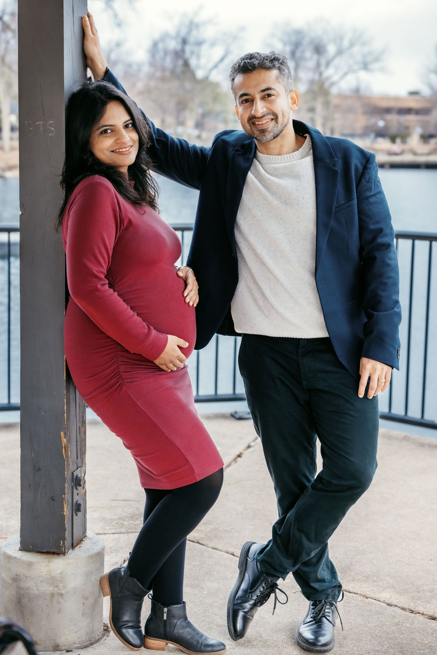 Expecting couple posing together beside a metal pole near a waterfront.