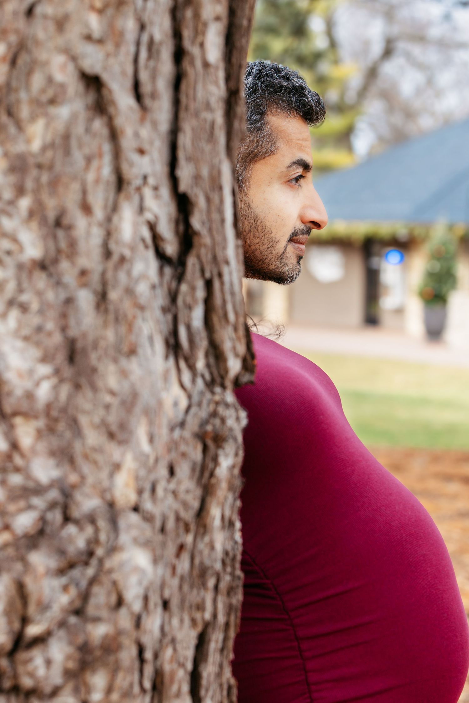 Side profile of pregnant woman in red dress beside a large tree outdoors.