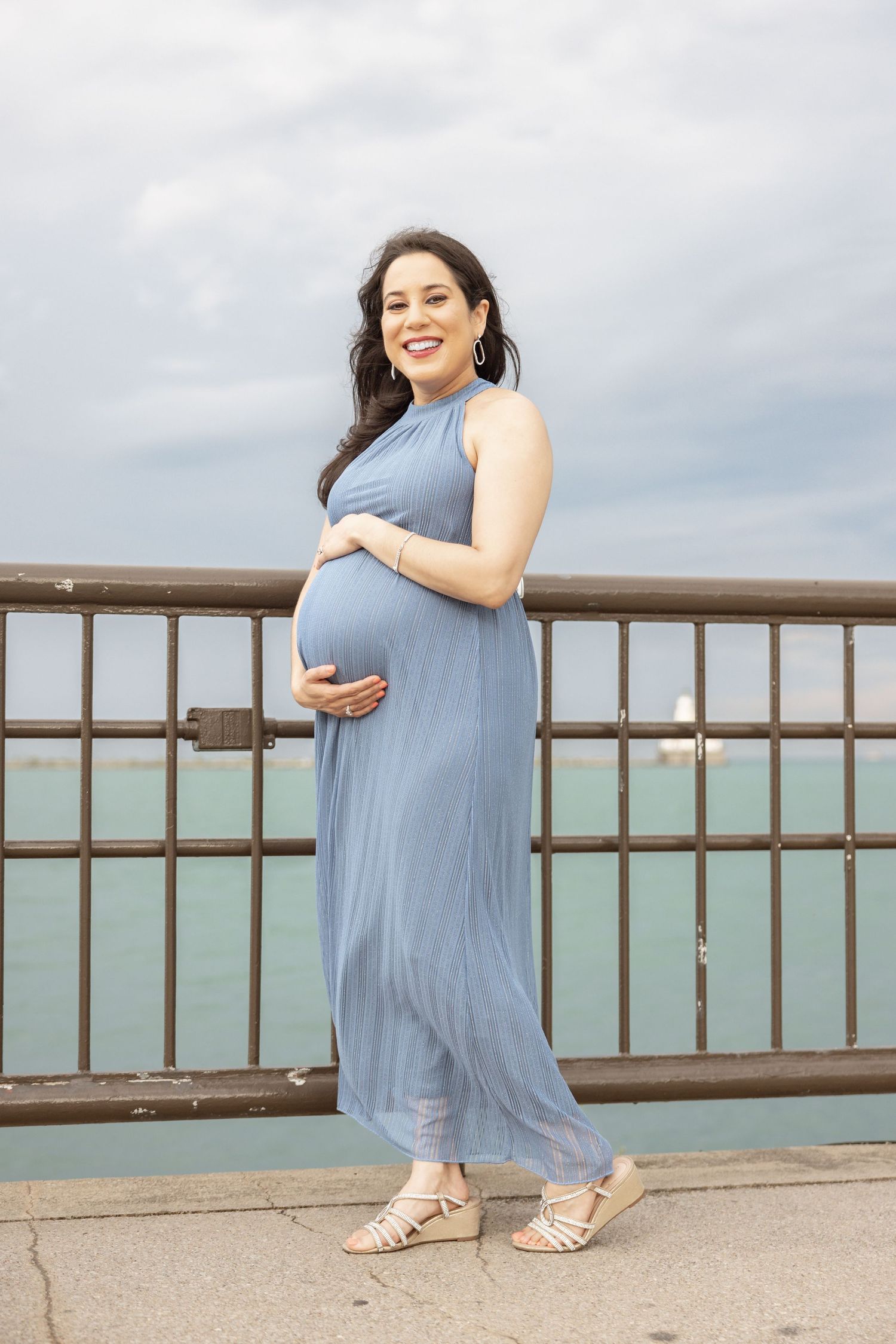 Happy pregnant woman in blue dress posing by metal railing near water.