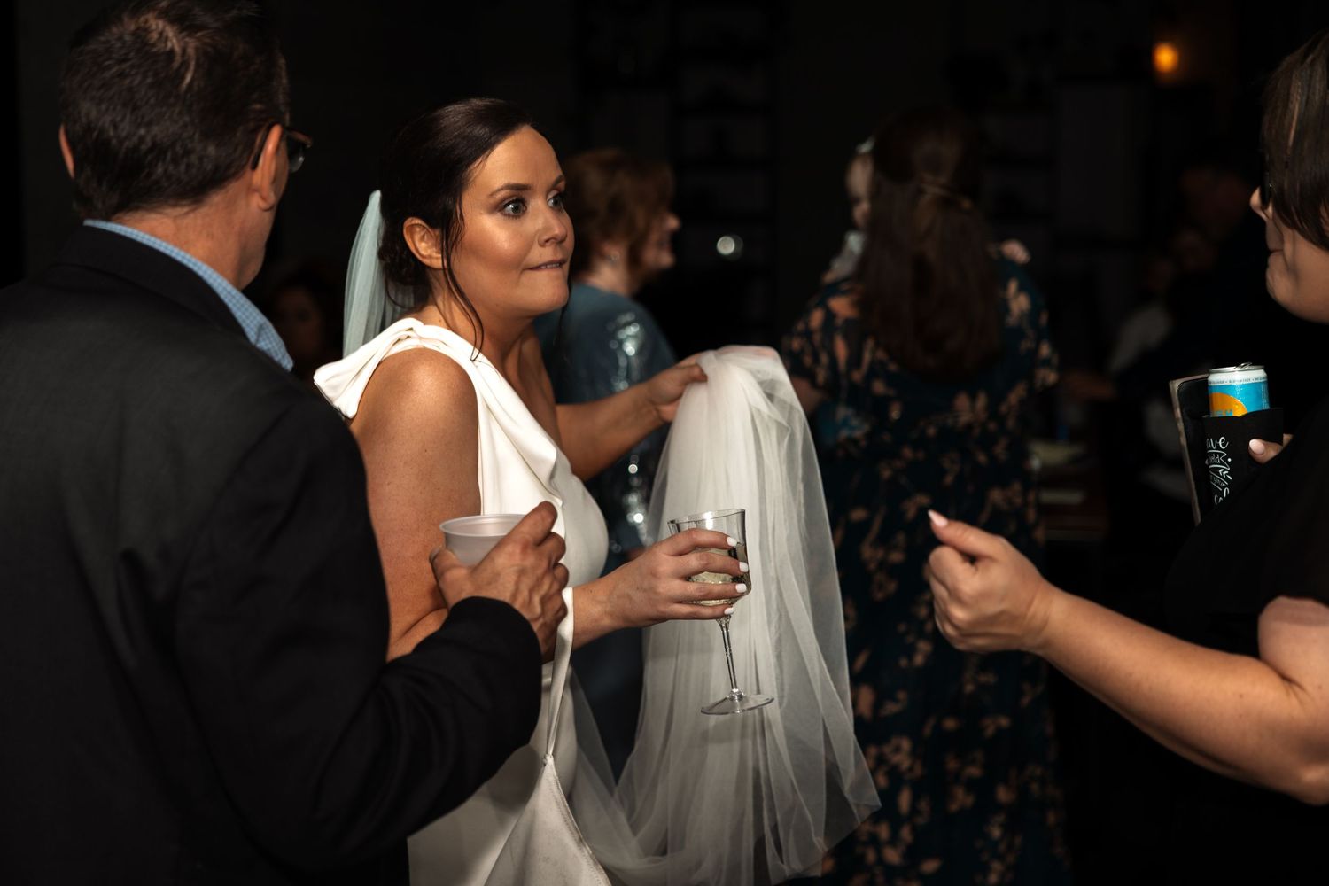 Bride in white gown holds champagne glass while mingling with guests at lively wedding reception.