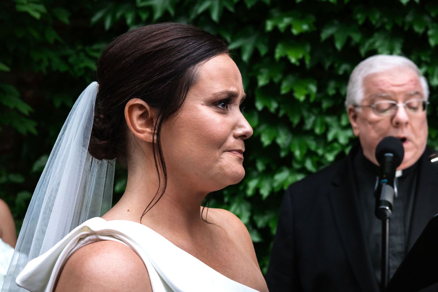 Emotional bride in white gown and veil listens to officiant during an outdoor wedding ceremony.