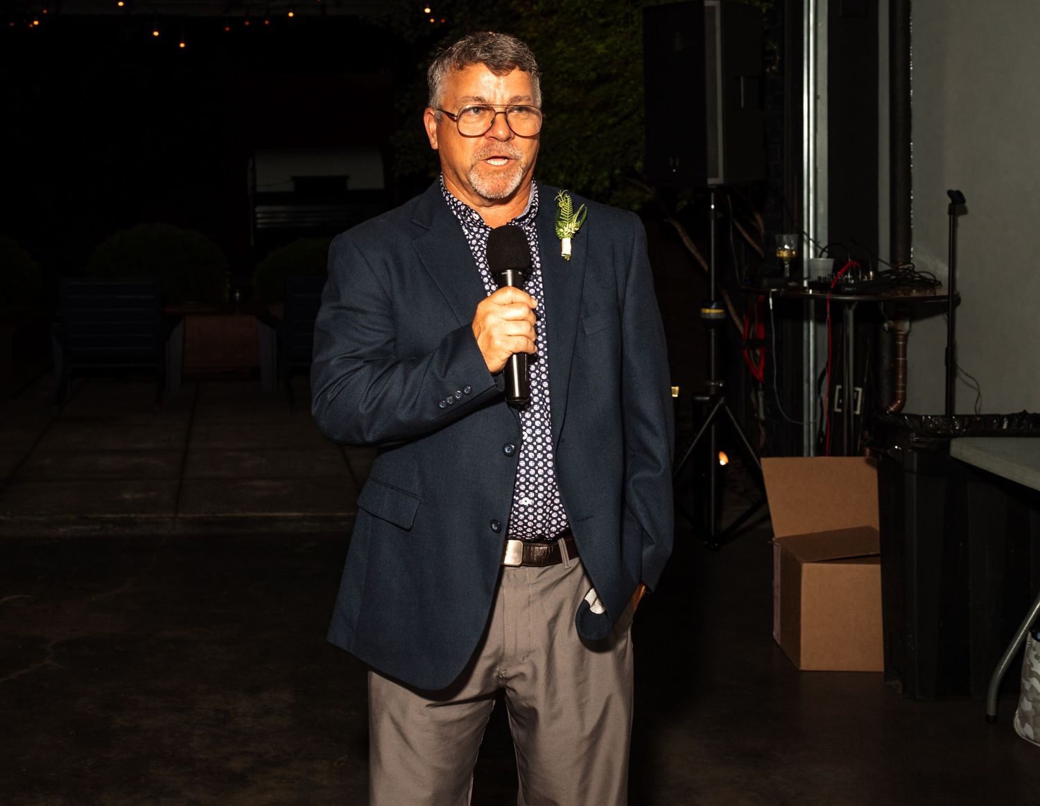 Man in navy blazer with boutonniere speaks into microphone while giving speech at outdoor wedding reception.