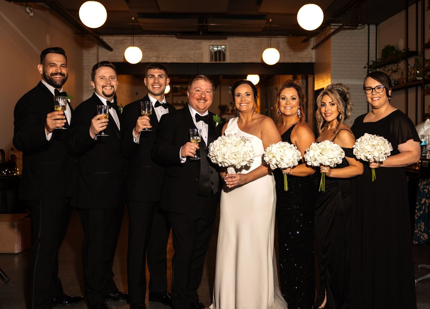 Elegant wedding party posing together with bride in white dress and bridesmaids holding white bouquets.