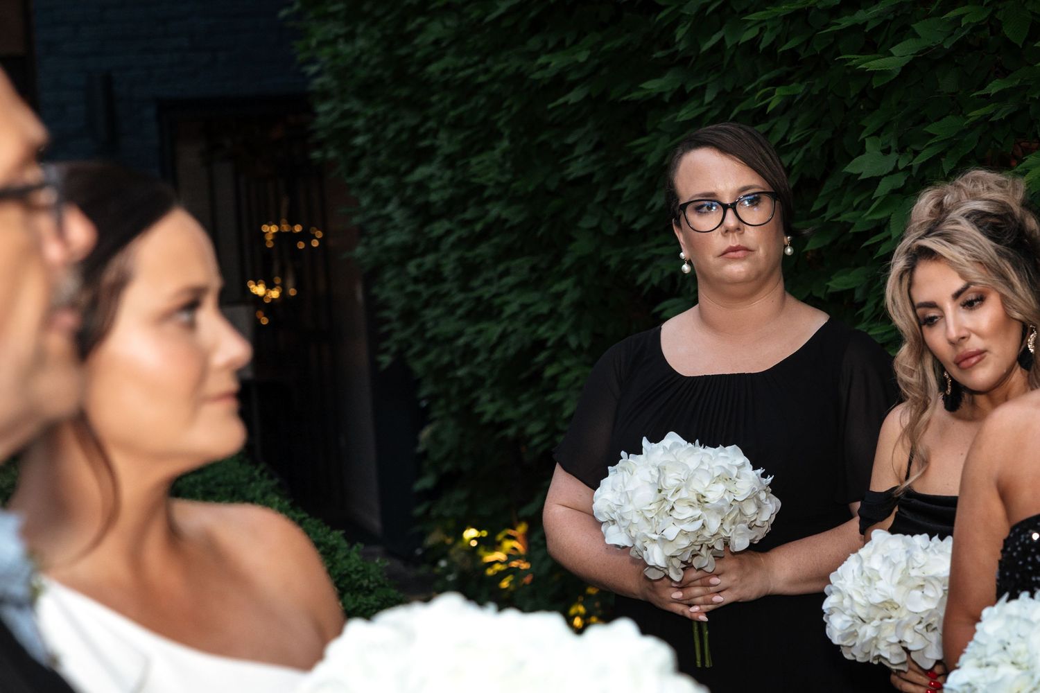 Bridesmaid in black dress with glasses holding white hydrangea bouquet watching outdoor wedding ceremony.