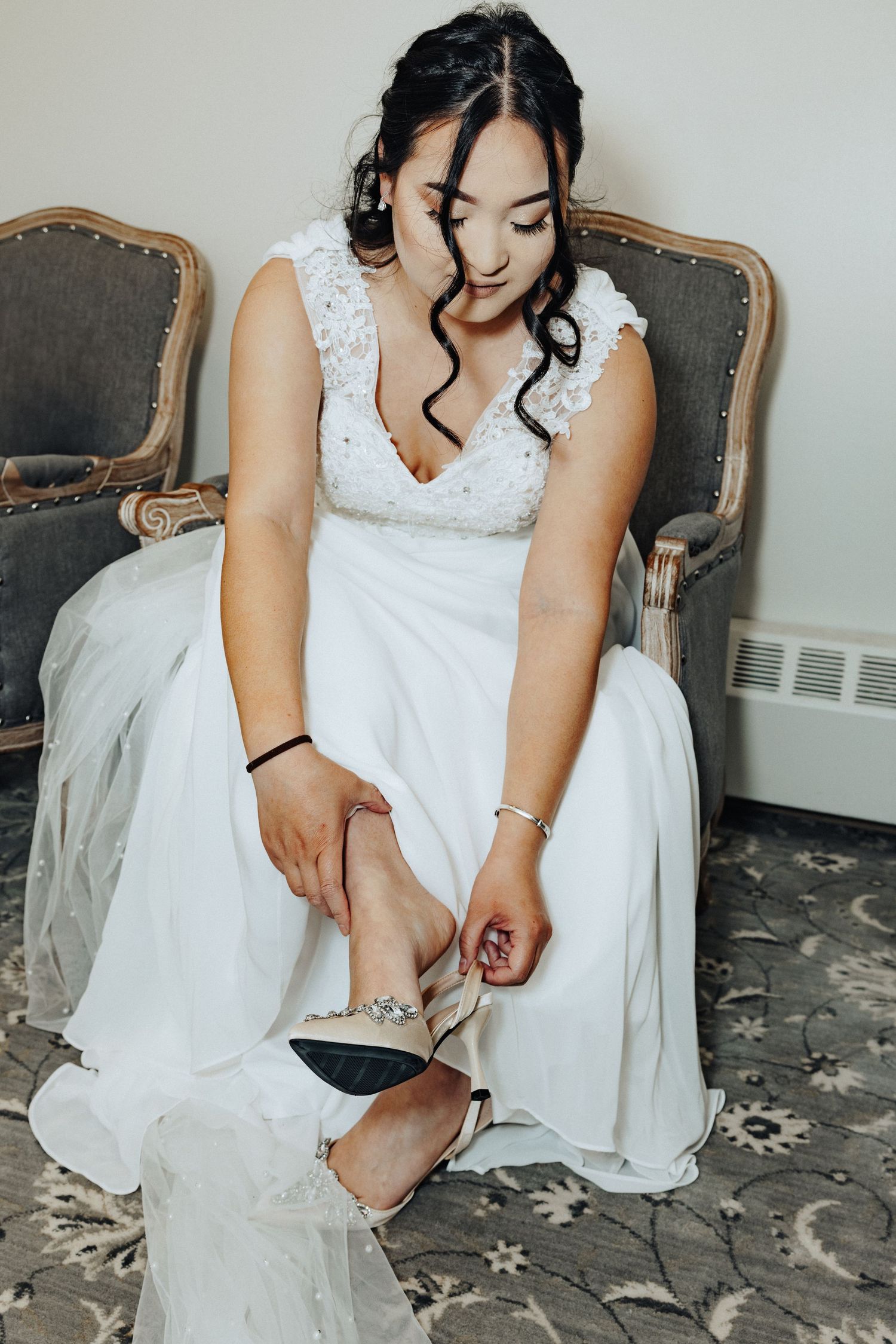 Bride in white lace wedding dress sitting on vintage chair adjusting her embellished heels.