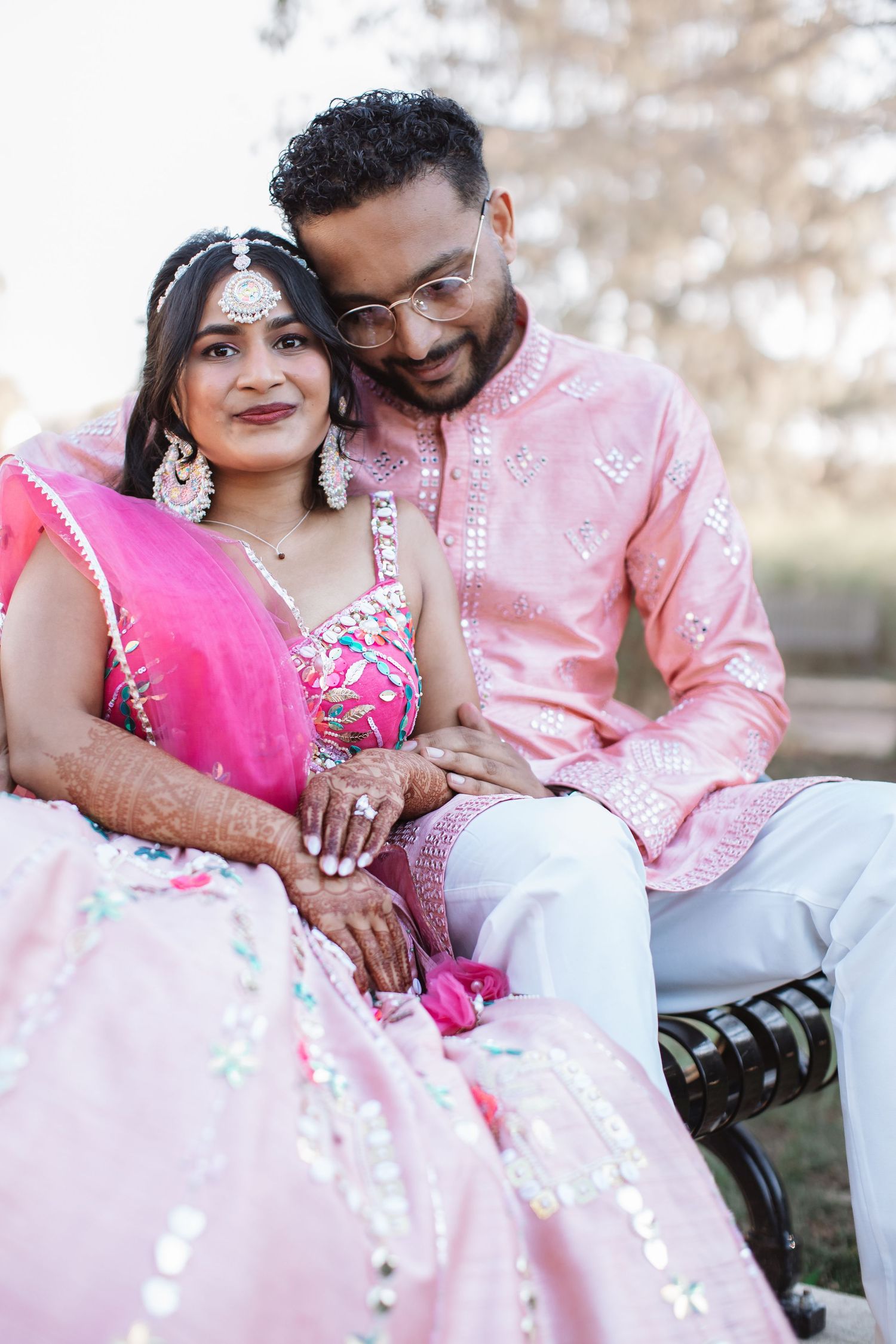 Indian couple in pink traditional wedding attire sitting closely together outdoors.