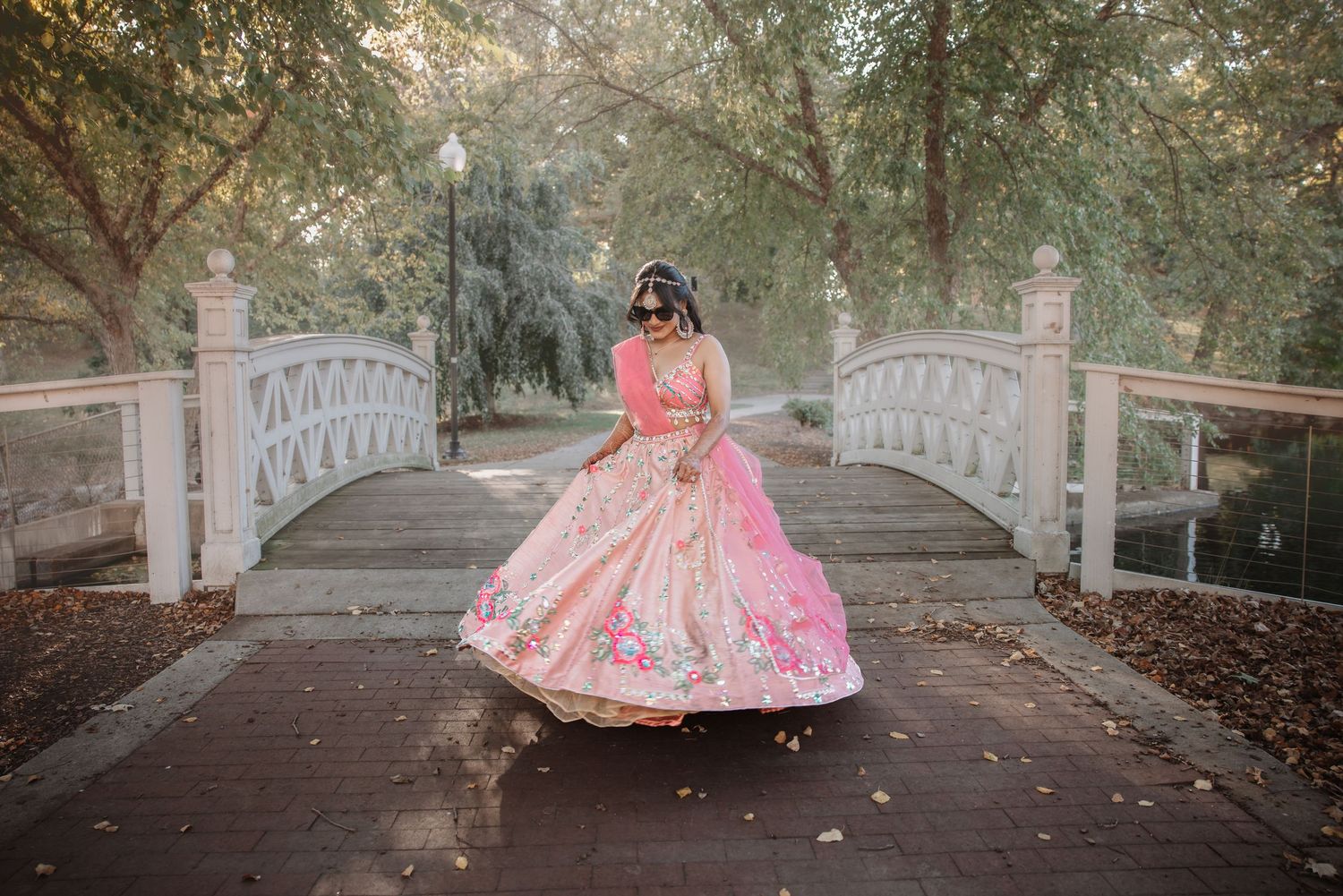 Indian bride spinning in elegant pink lehenga choli near ornate white bridge outdoors.