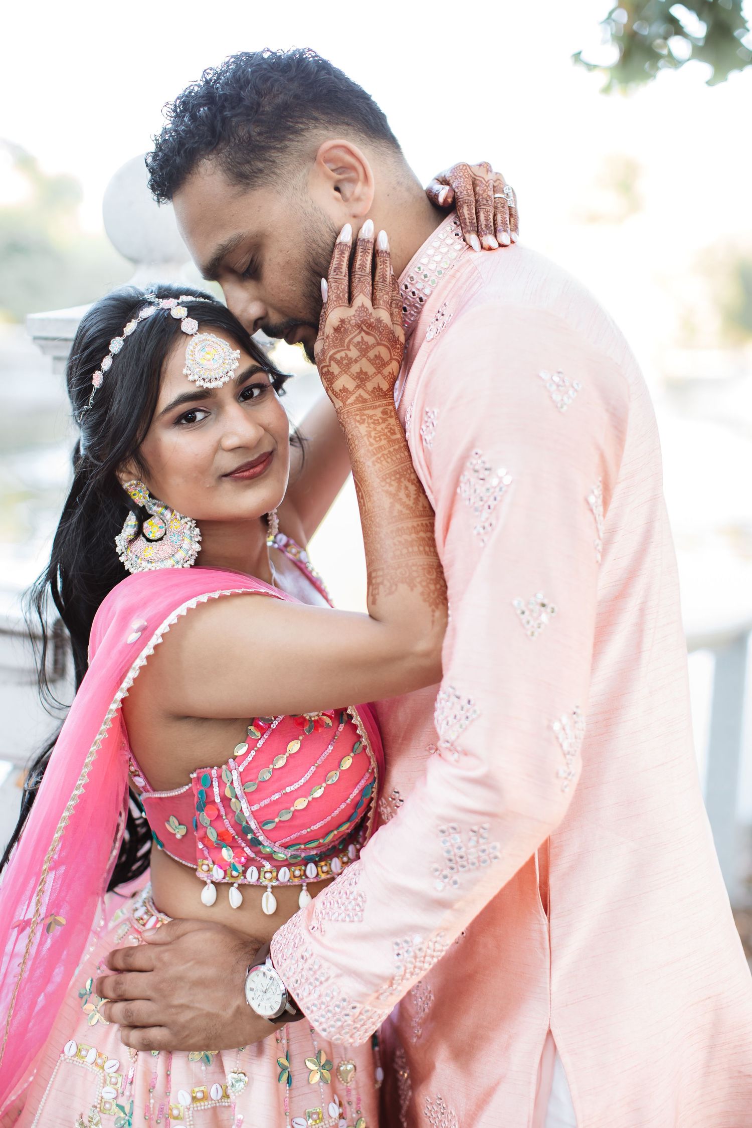 Indian bride in pink lehenga smiling as groom in peach kurta kisses her henna-covered hand outdoors.
