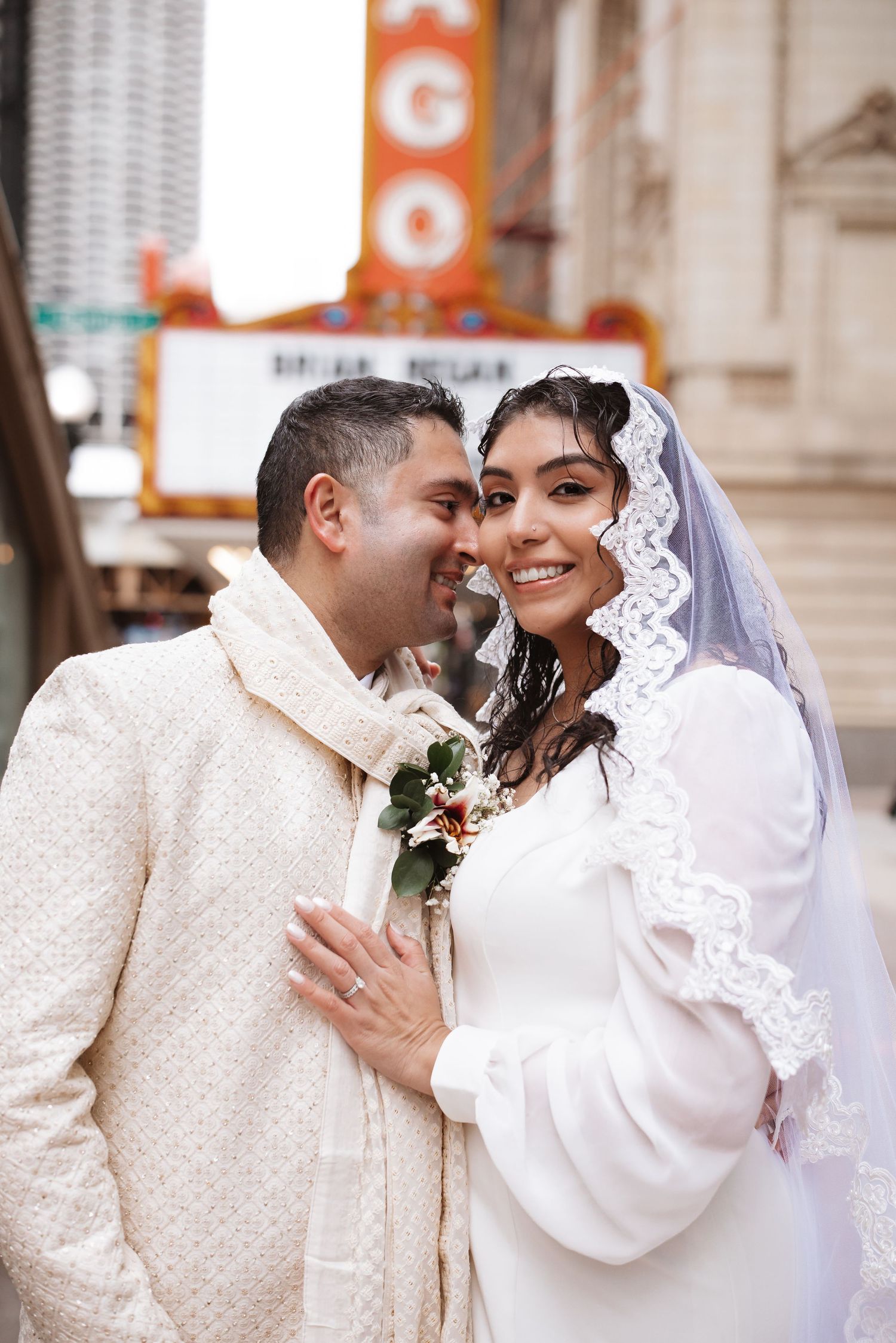 Smiling South Asian bride in lace veil embraces groom in cream sherwani outside Chicago Theatre.