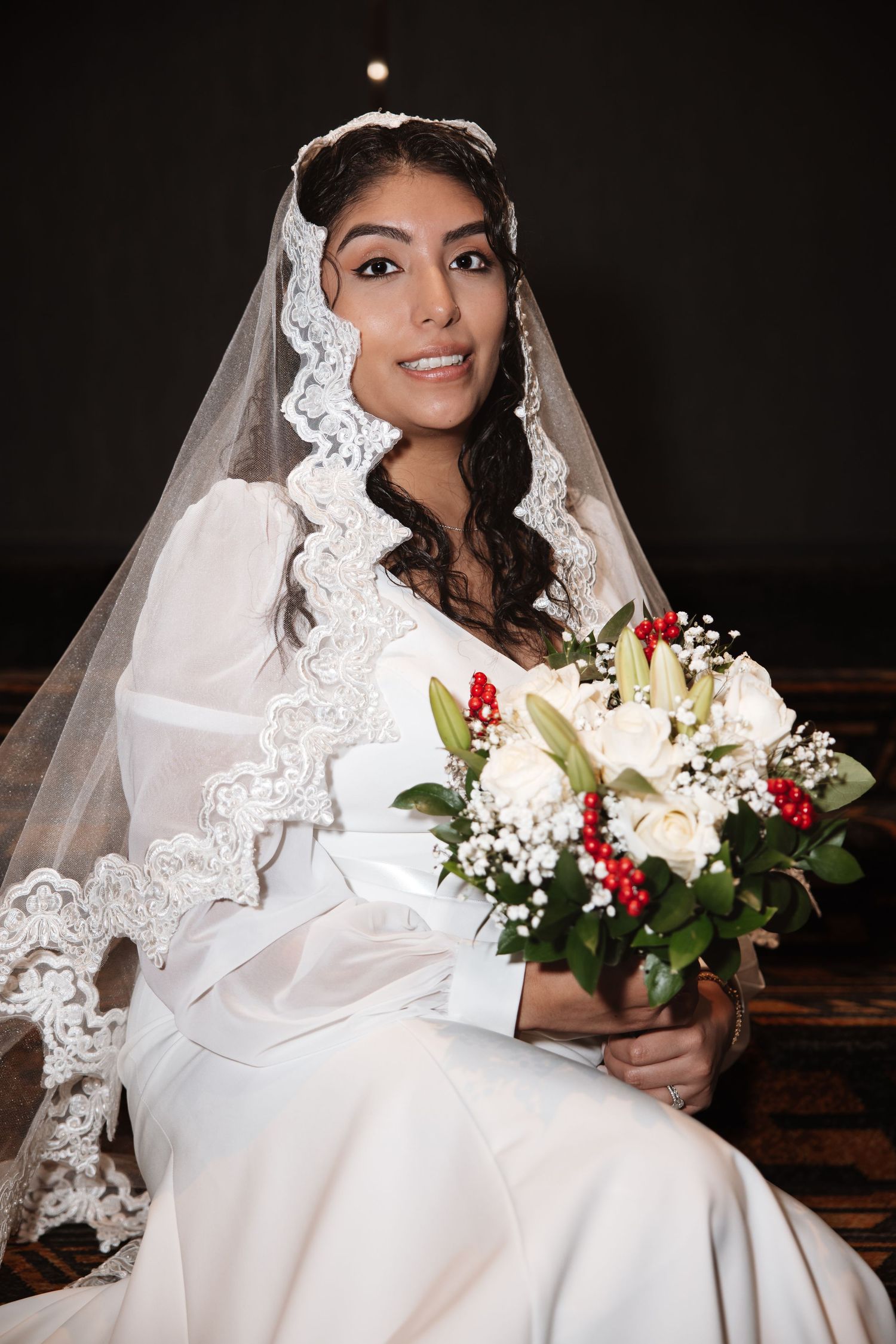 Seated bride in white gown and lace veil smiles while holding bouquet of white roses and red berries.