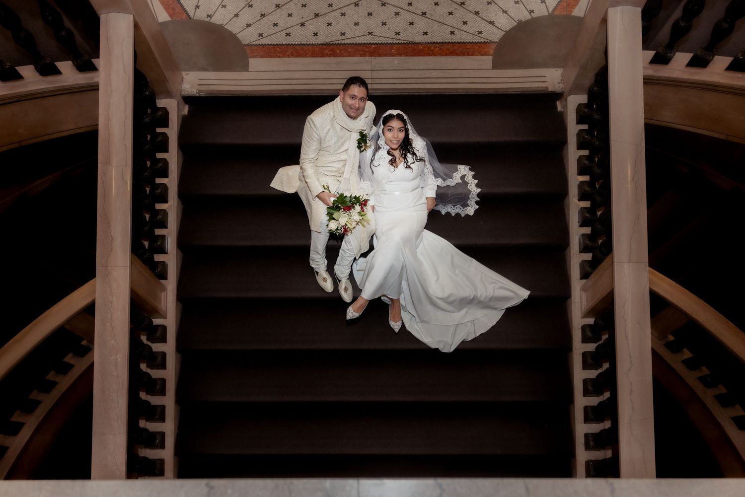 Aerial view of bride and groom sitting on wooden spiral staircase, bride in white gown holding bouquet.