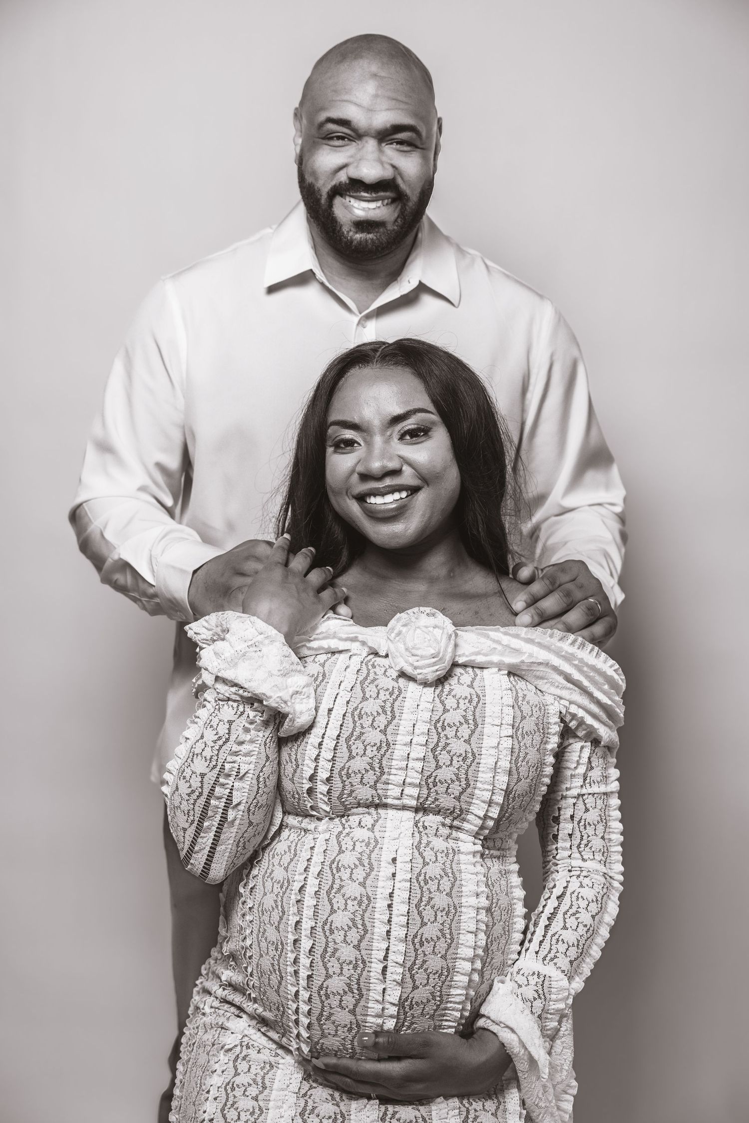 Black and white portrait of smiling pregnant couple, woman in lace dress holding baby bump.