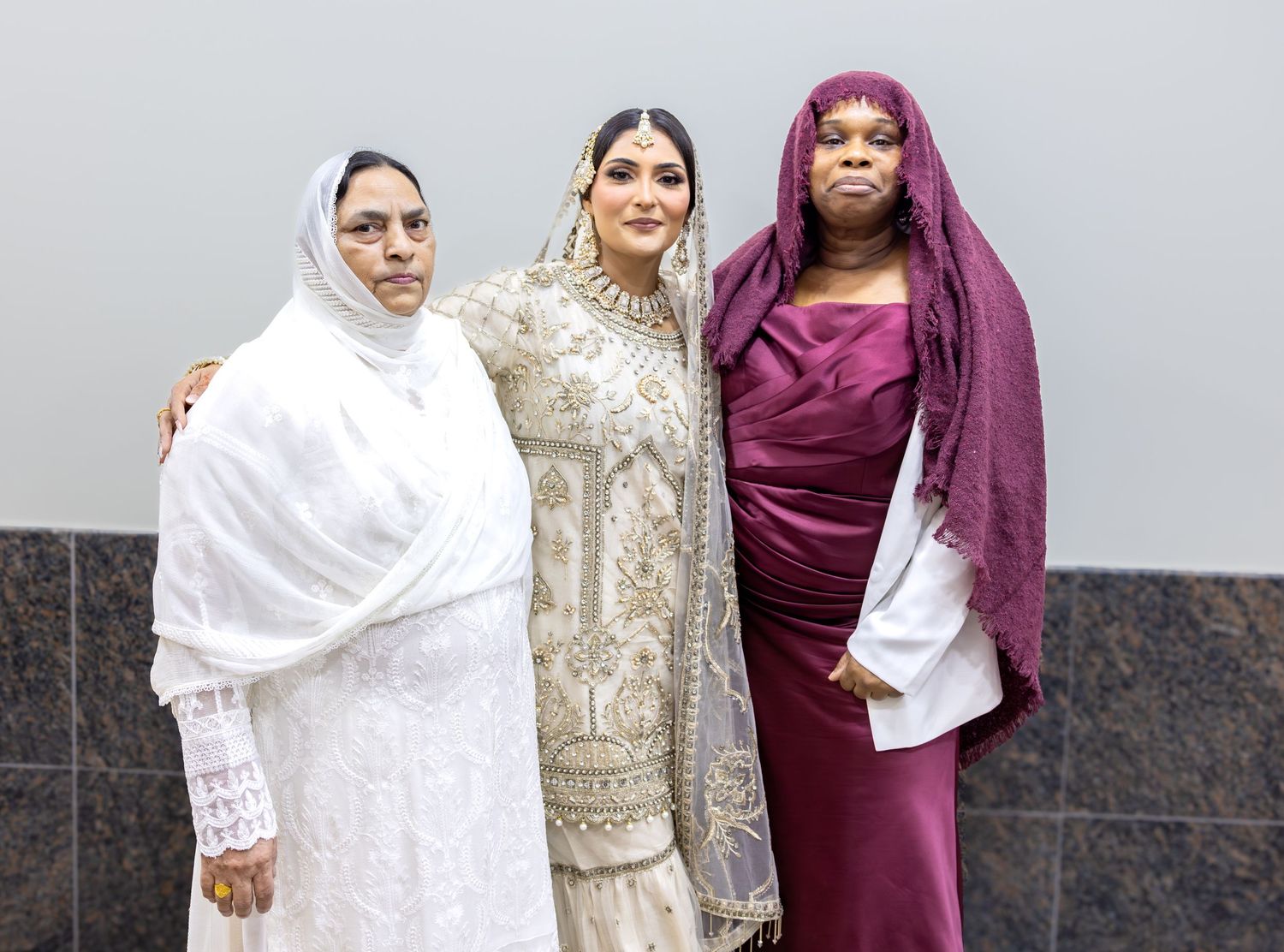 Three women pose together, with a bride in white bridal attire flanked by two women in white and burgundy hijabs.