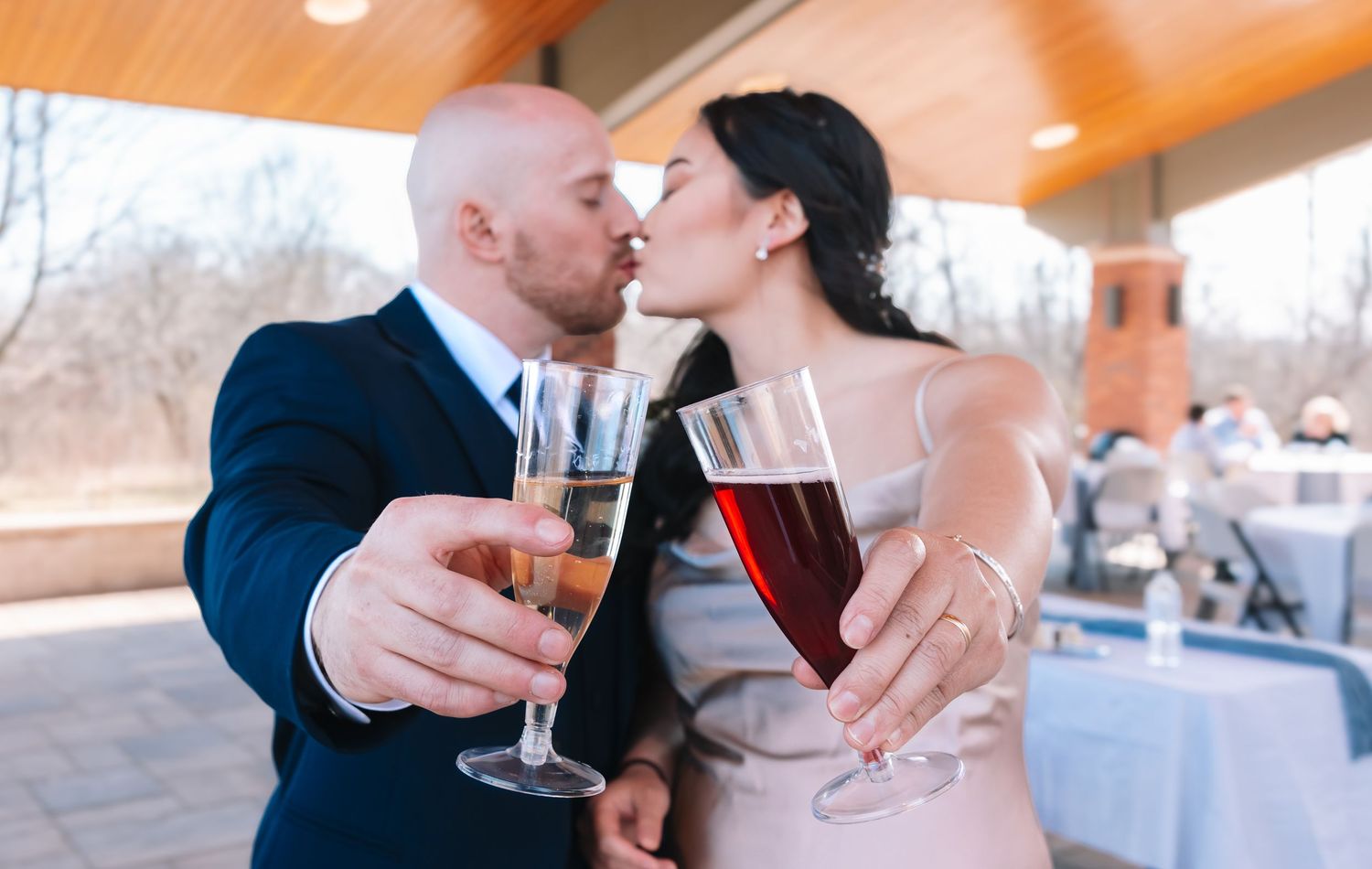 A couple shares a kiss while toasting with champagne and red wine at their outdoor wedding reception.