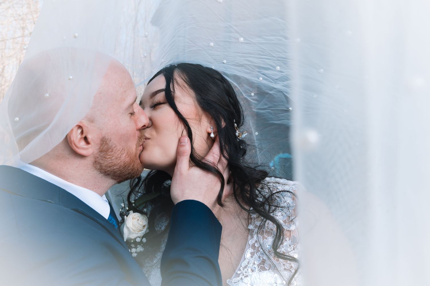 Groom tenderly kisses bride while cradling her face beneath a flowing wedding veil outdoors.