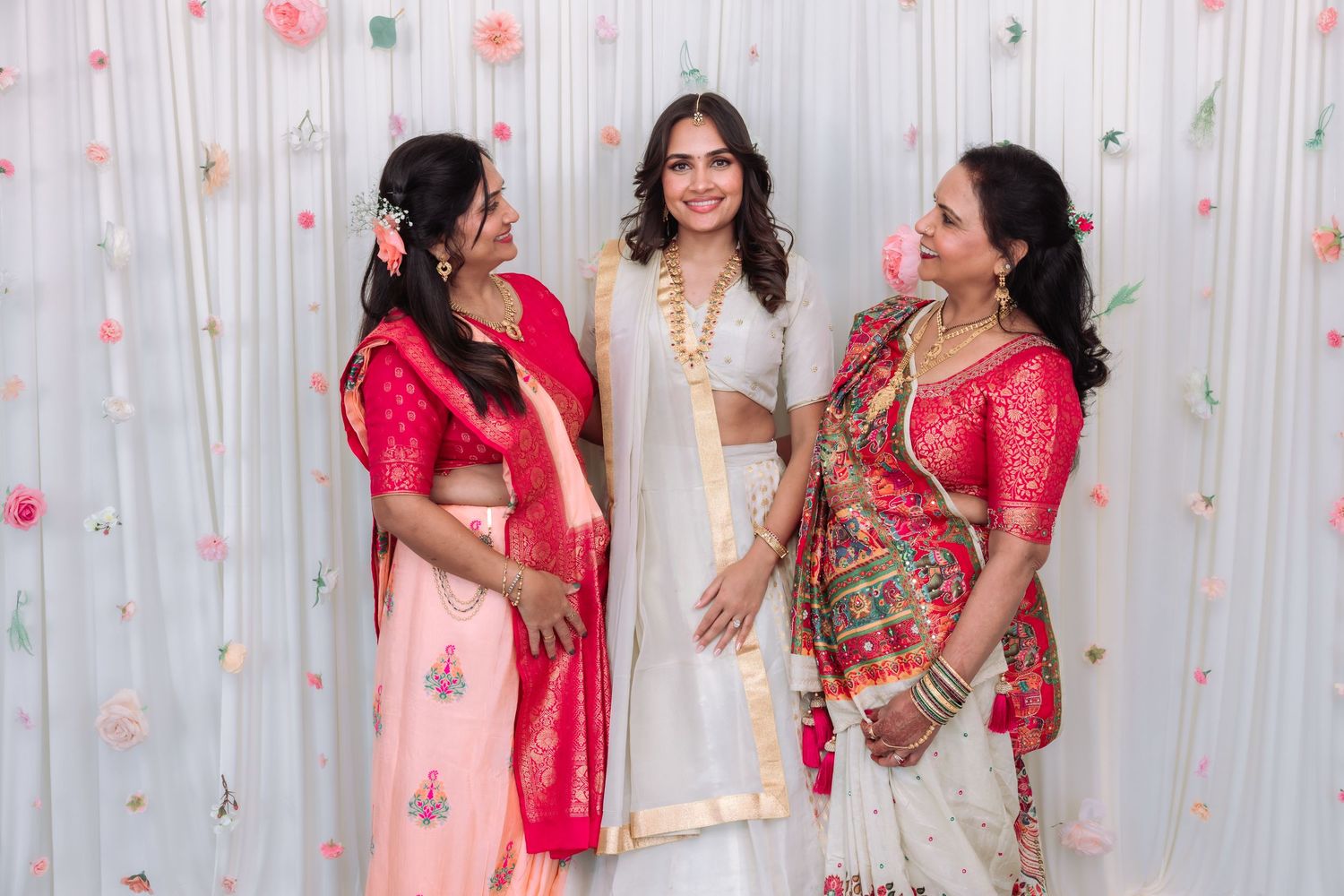 Three Indian women laughing together in traditional pink and white wedding outfits.