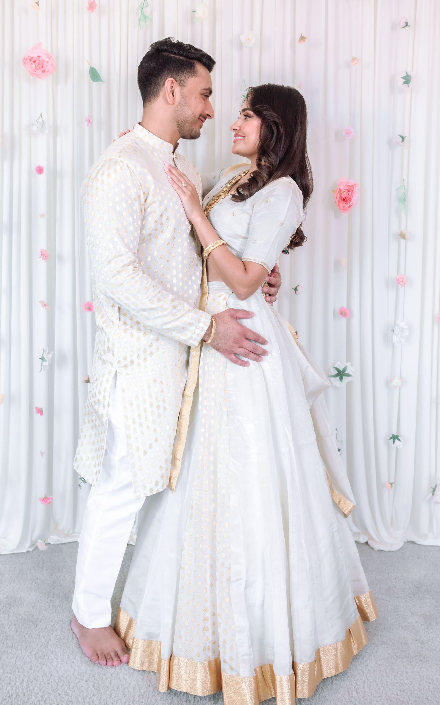 Indian couple in matching white and gold traditional outfits embrace at a floral decorated ceremony.
