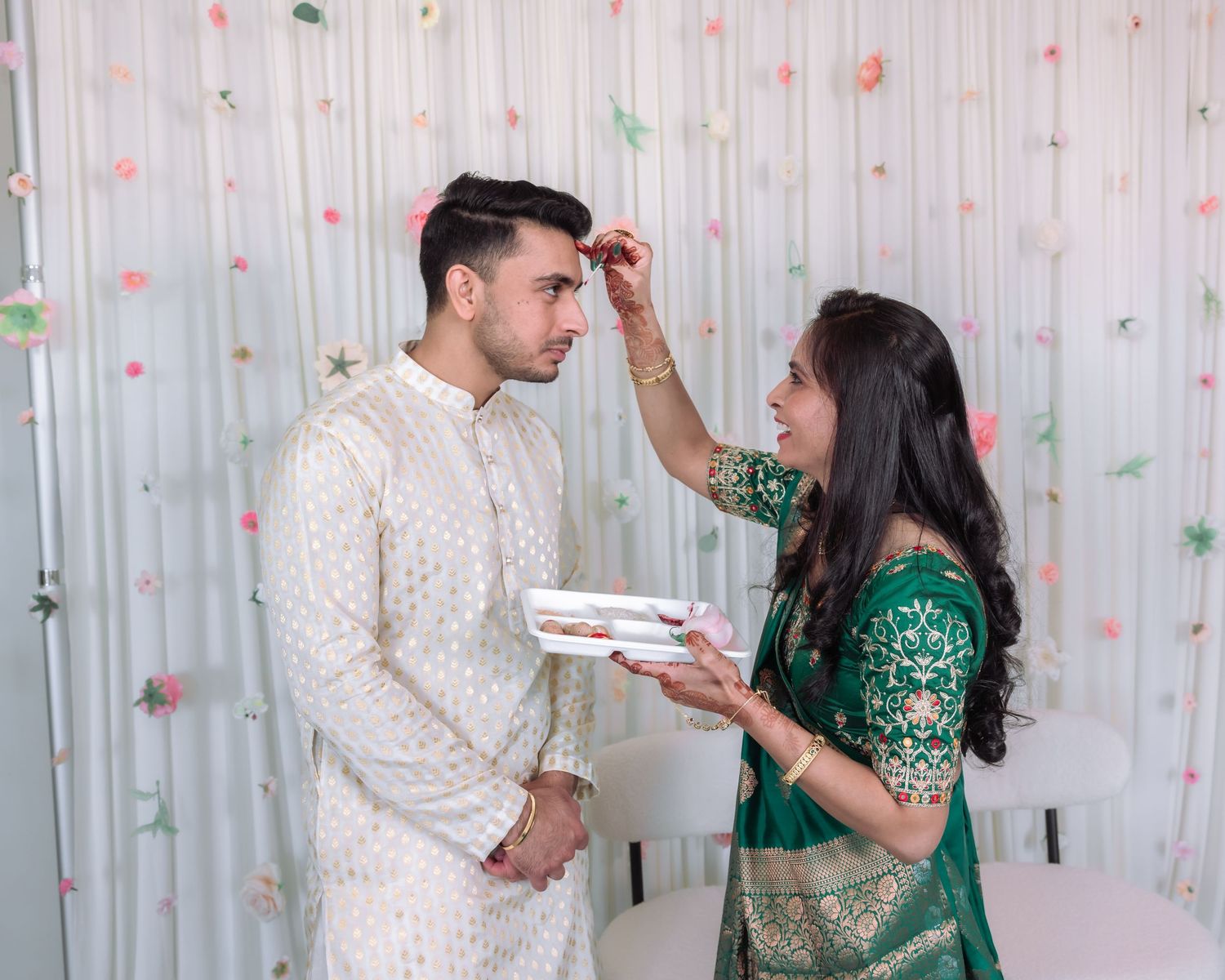 Woman in green saree applying tilak to groom in white sherwani during Indian wedding ritual ceremony.