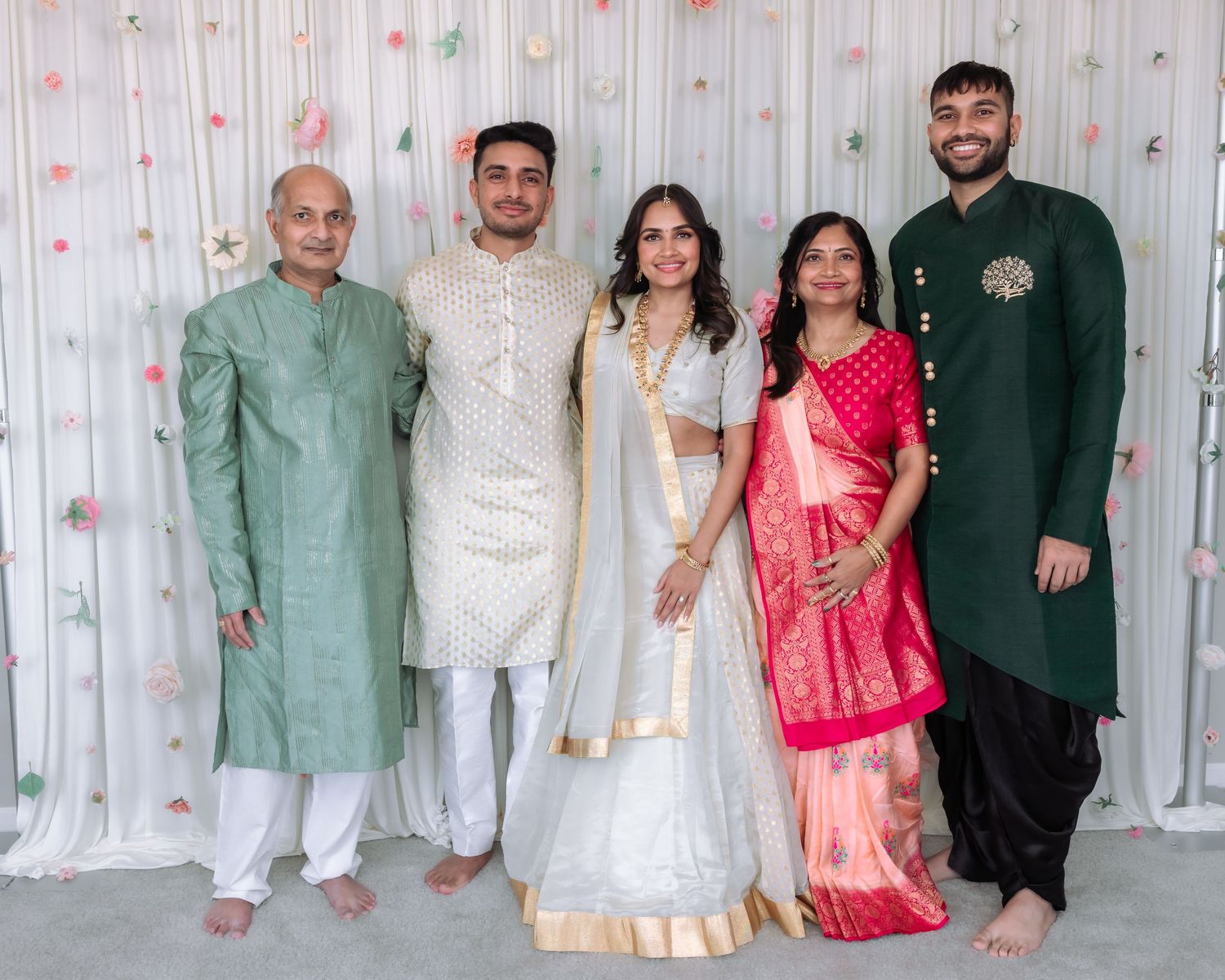 Five family members in traditional Indian wedding attire pose together against a floral backdrop.
