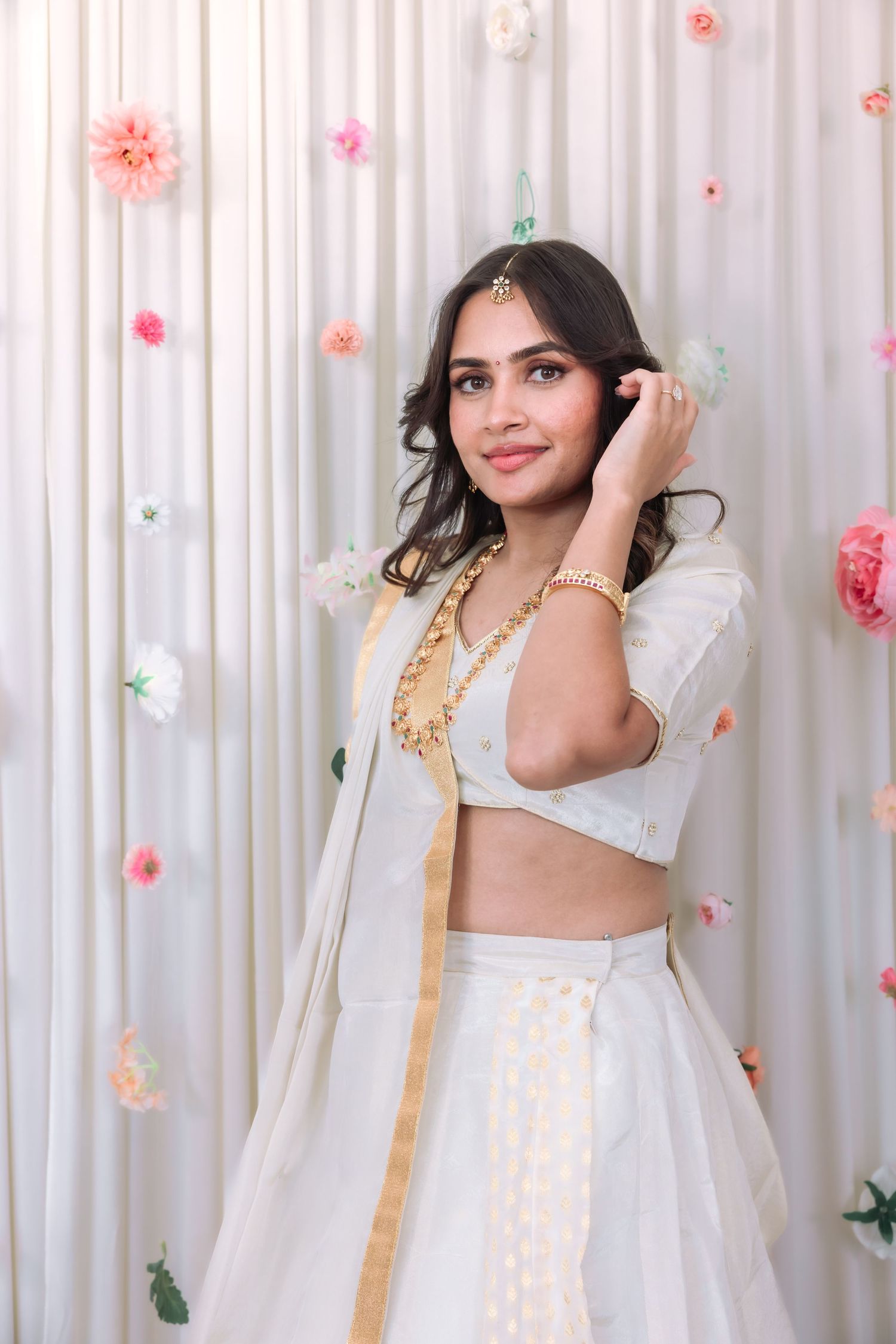 Young woman in white lehenga with gold accents and maang tikka posing before floral backdrop.
