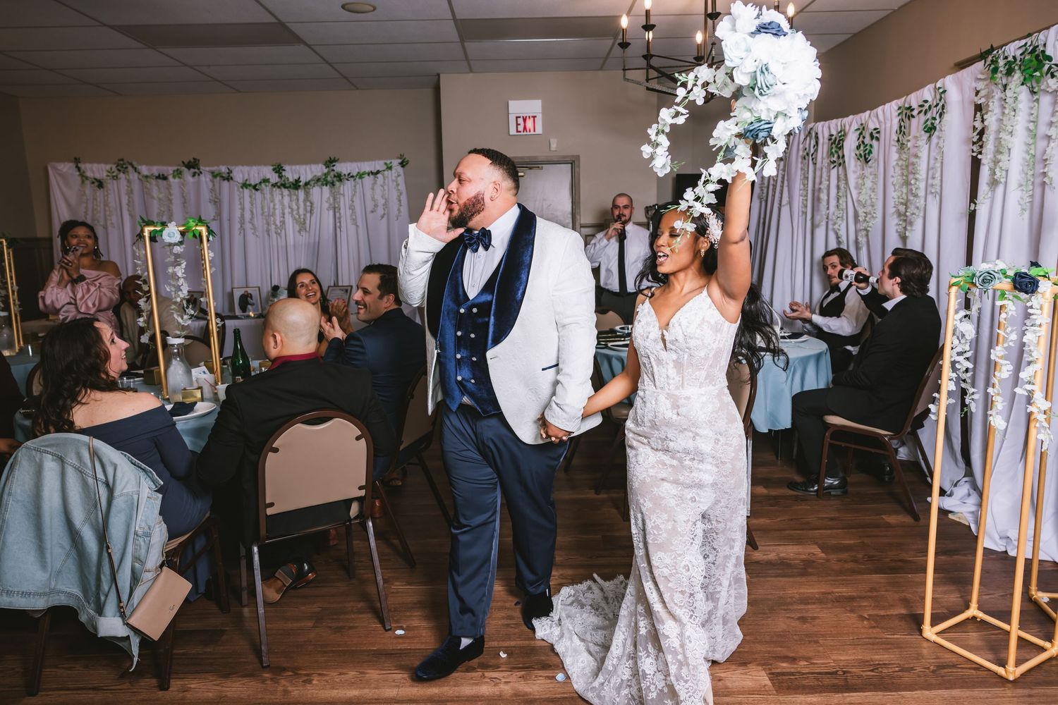 Bride in lace gown and groom in white tuxedo making grand entrance at wedding reception.