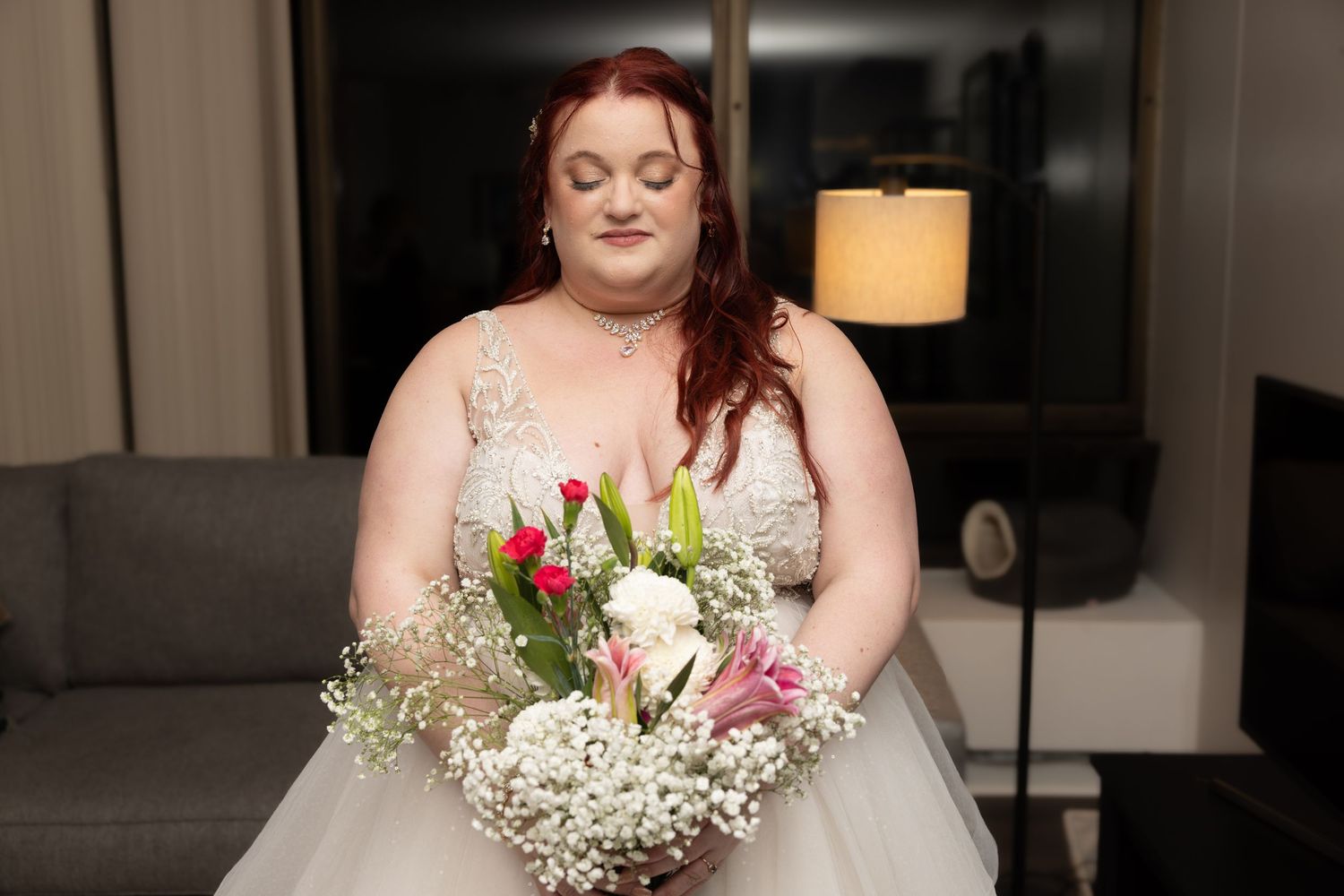 Red-haired bride in elegant wedding gown looks down at vibrant bouquet in softly lit room.